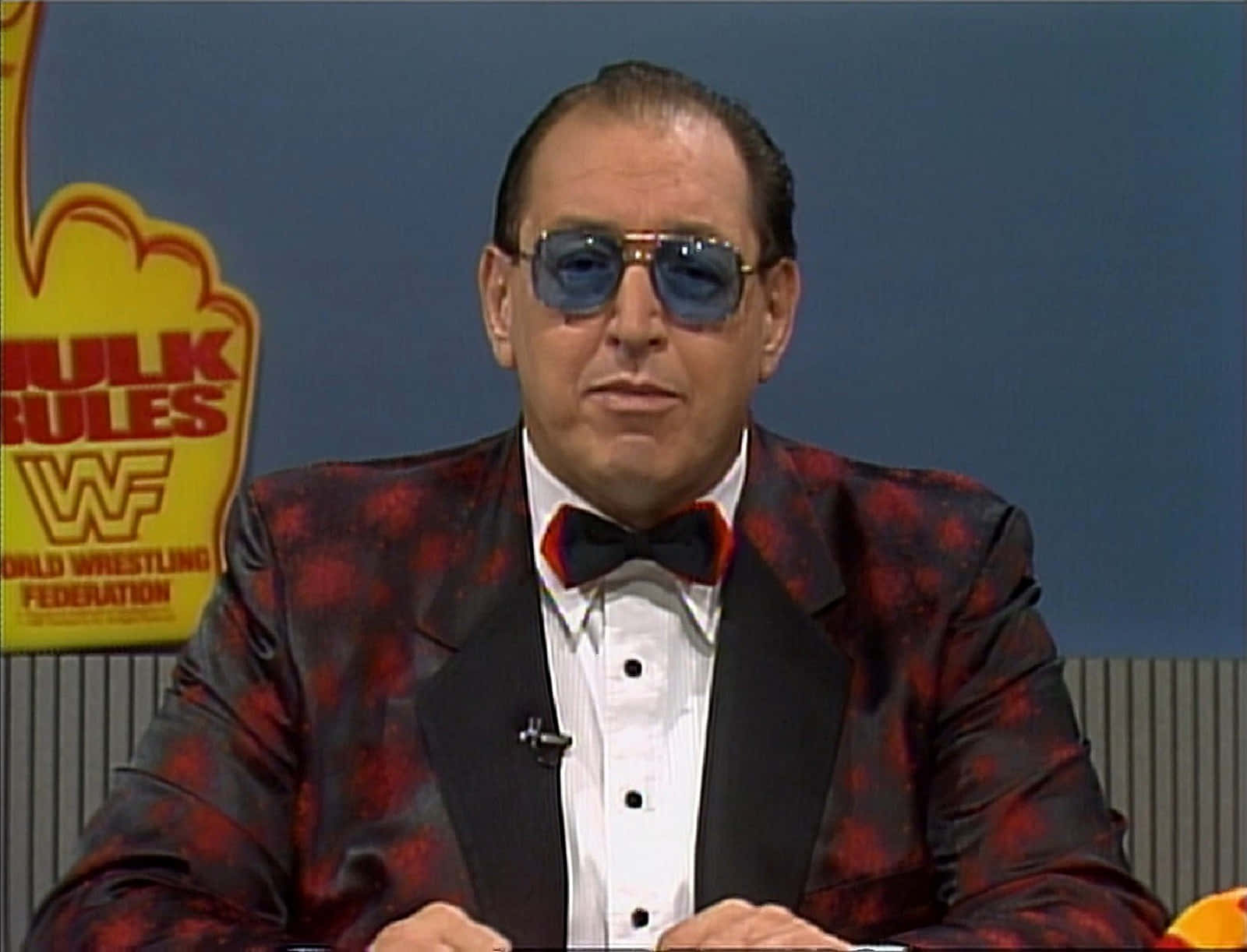 Gorilla Monsoon 桌布 壁紙 — 一名身穿紅色圖案燕尾服和太陽眼鏡的男子坐在一張後面有「WWE」標誌的辦公桌前。