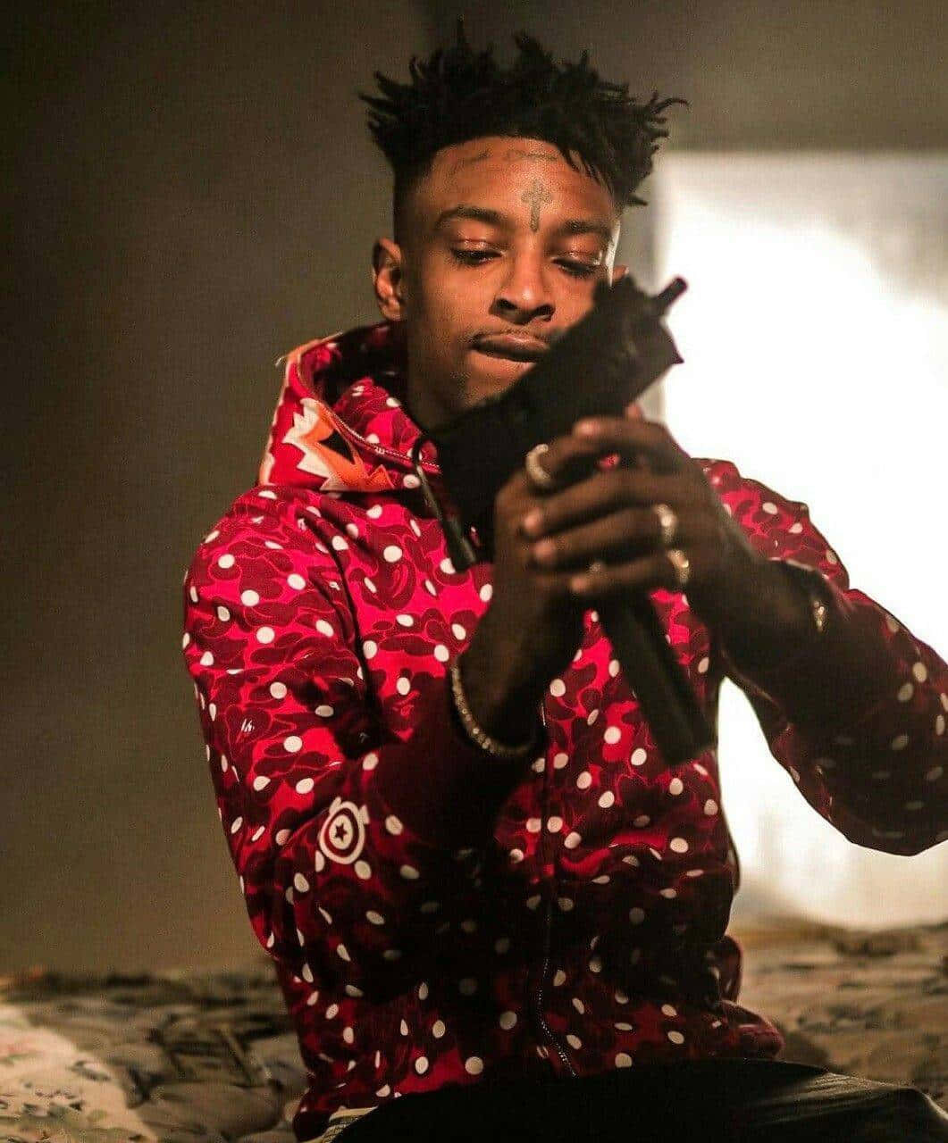 21 Savage 桌布 壁紙 — 一名身穿紅色圖案連帽衫的男子在戲劇性燈光下持槍，表情嚴肅。