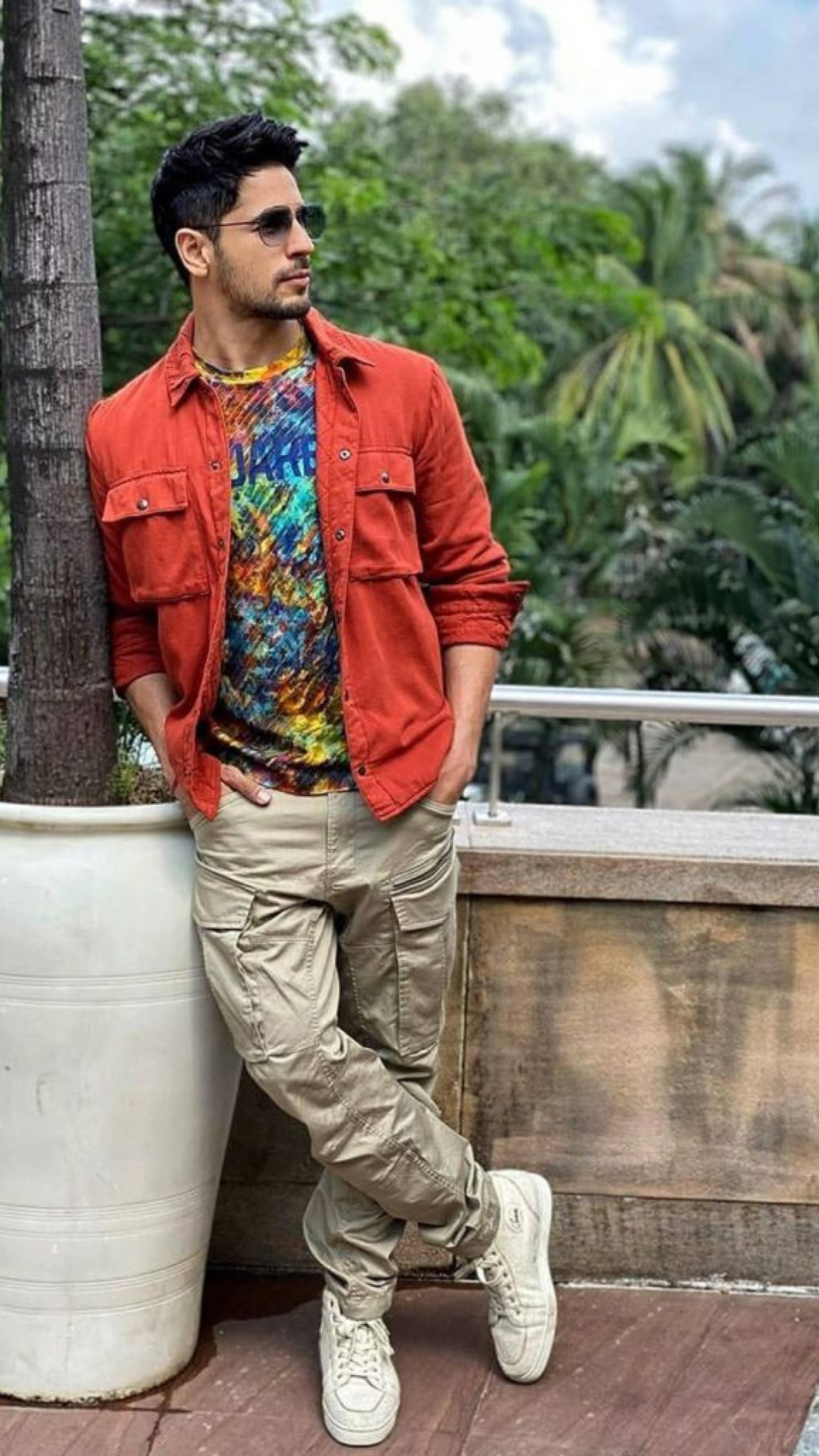 Sidharth Malhotra 桌布 壁紙 — 一名身穿紅色外套和彩色襯衫的男子，在一個被茂密綠色棕櫚樹環繞的陽台上，隨意地靠著一個大型白色花盆。