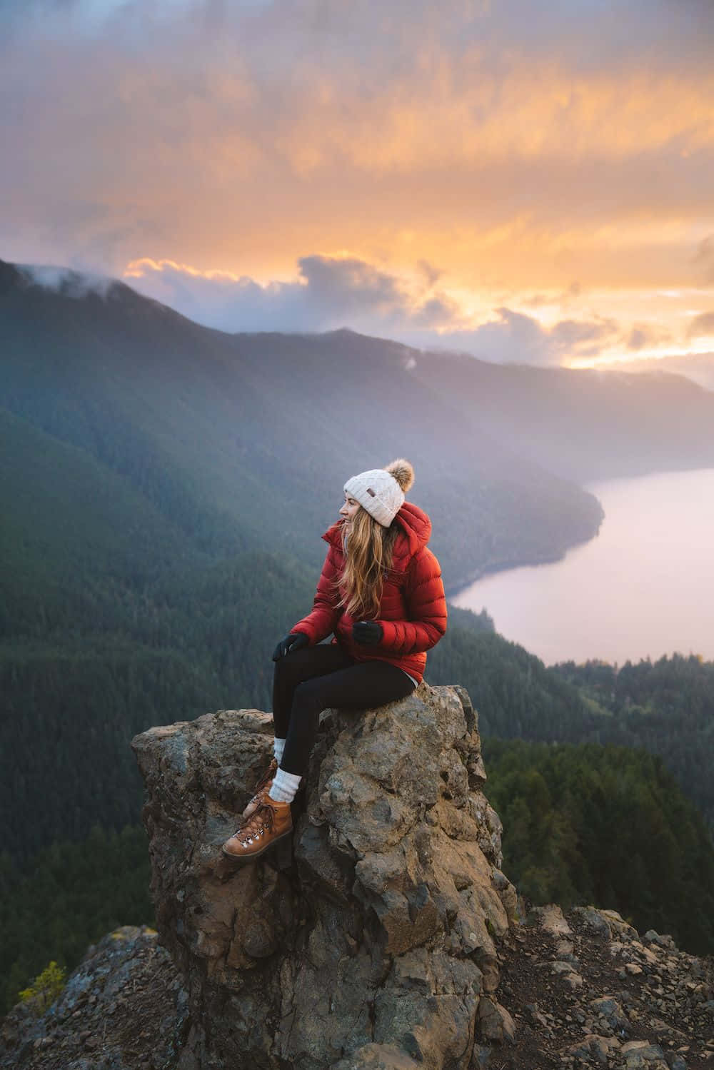 Winter Hiking 桌布 壁紙 — 一名身穿紅色外套和白色無邊帽的女子坐在岩石峭壁上，俯瞰日落時分薄霧籠罩的山谷。