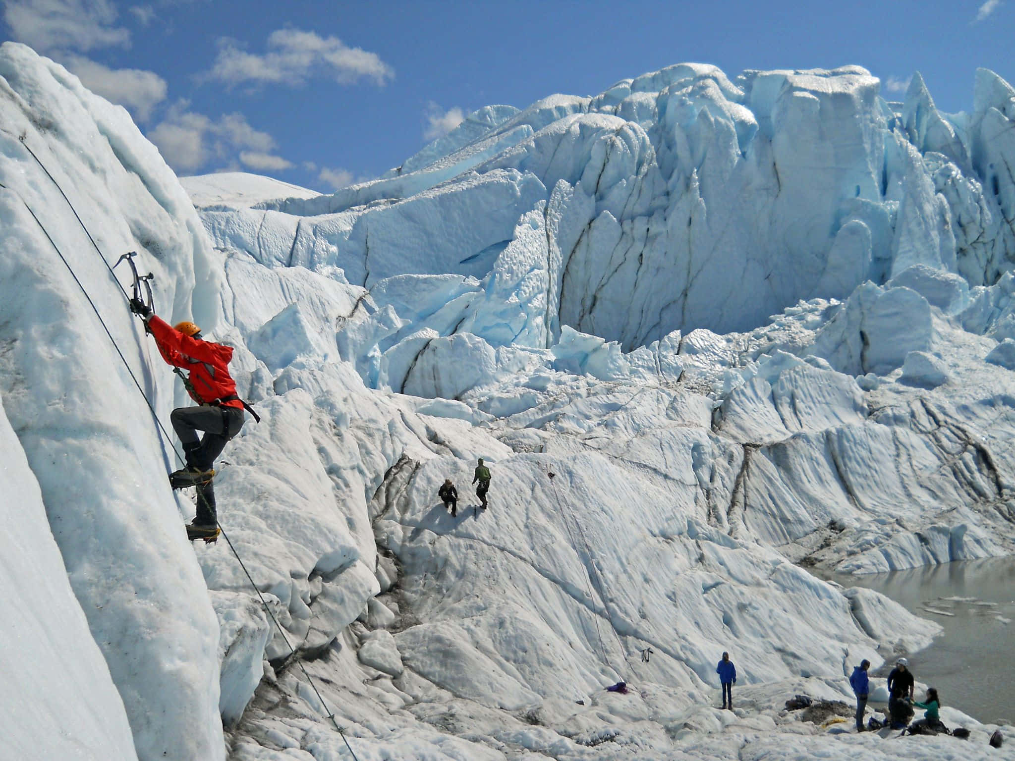 Ice Climbing 桌布 壁紙 — 一名身穿紅色外套的攀岩者在明亮的藍天下攀登巨大的冰川，而其他人在下方徒步。