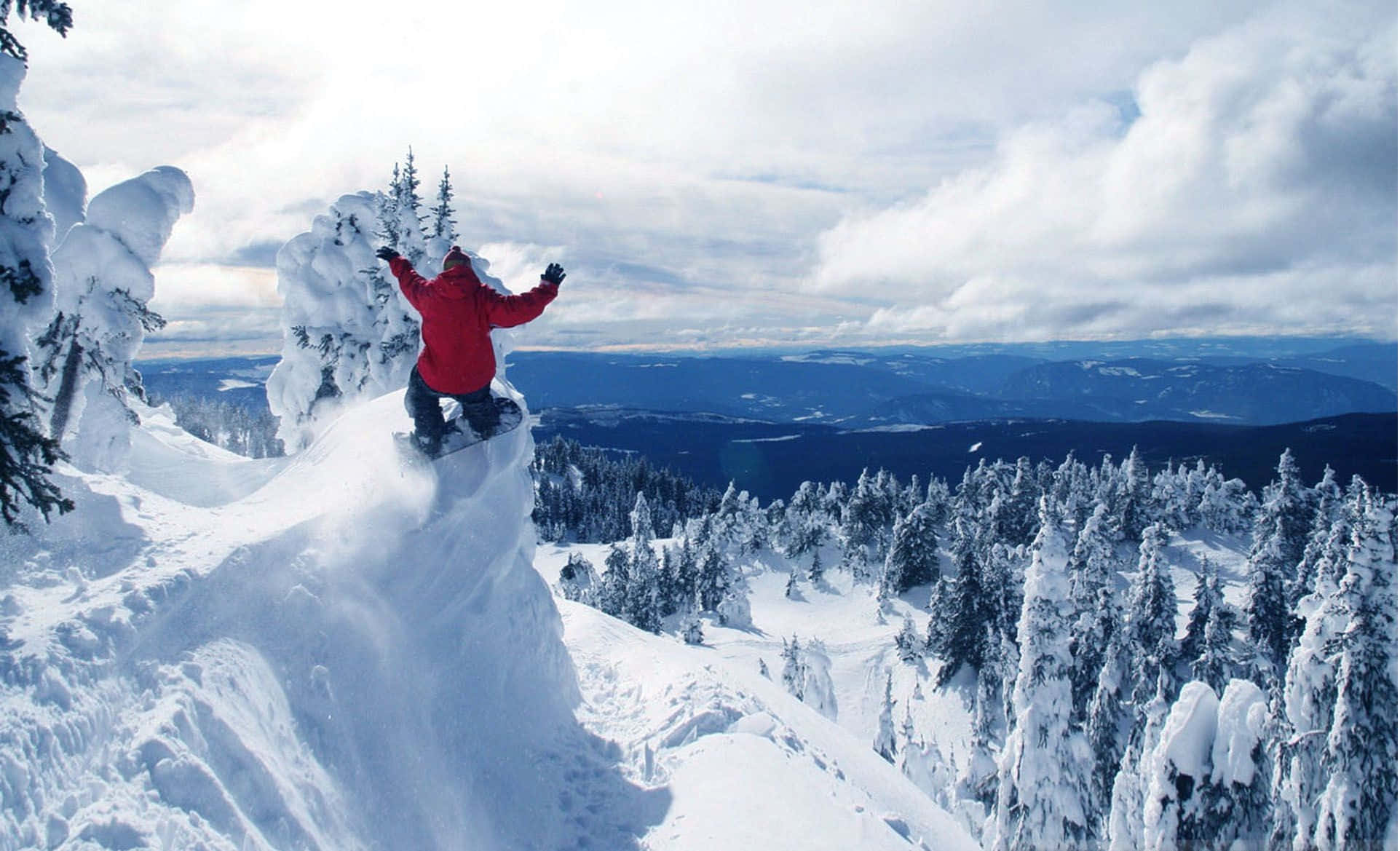 Winter Sports 桌布 壁紙 — 一名身穿紅色外套的滑雪板運動員從白雪覆蓋的懸崖上躍下，俯瞰著陰天下一片廣闊的冬季森林。