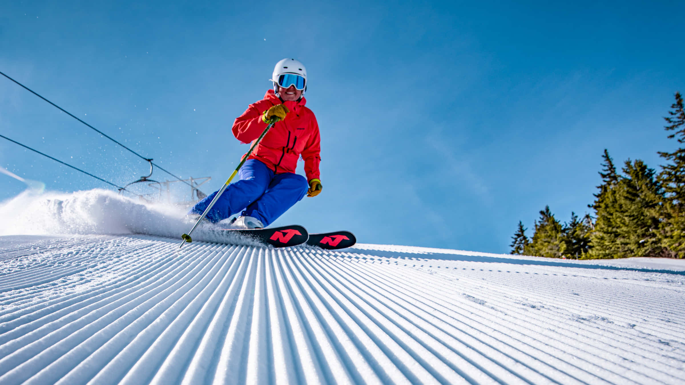 ski 桌布 壁紙 — 一名身穿紅色外套的滑雪者，在晴朗藍天下沿著整理過的雪坡滑下，背景有常綠樹。