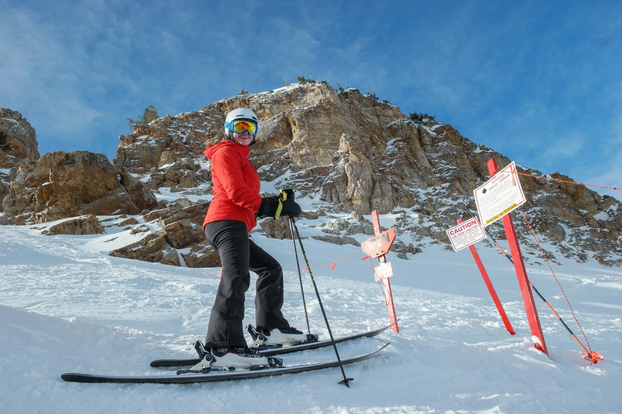 ski 桌布 壁紙 — 一名身穿紅色外套的滑雪者在雪坡上微笑，背景是岩石峭壁和藍天。