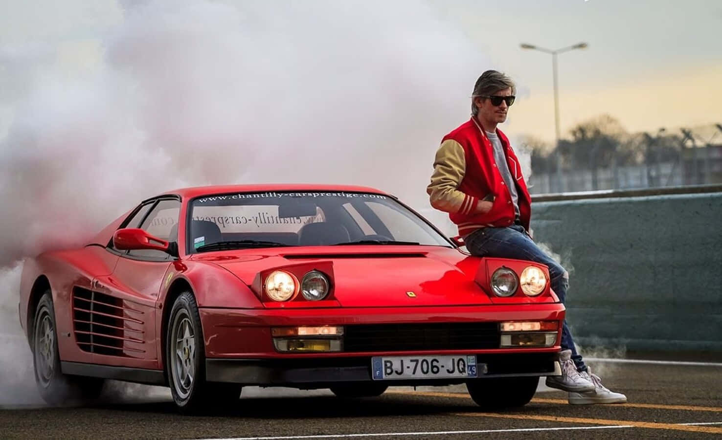 Ferrari Testarossa 桌布 壁紙 — 一名身穿紅色外套的男子自信地坐在一輛紅色法拉利上，在賽道上進行燒胎，煙霧滾滾。