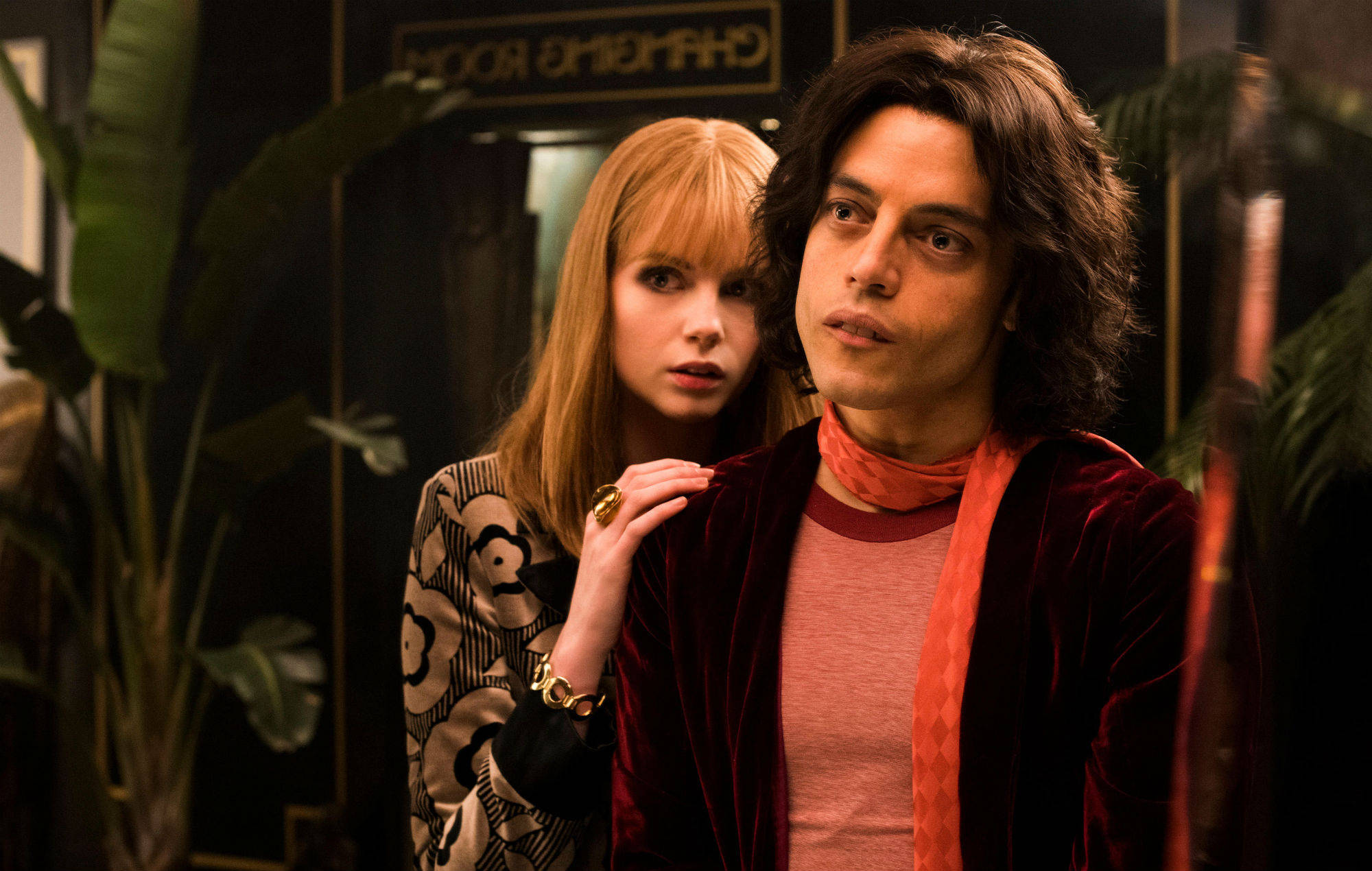 Rami Malek 桌布 壁紙 — 一名身穿紅色天鵝絨外套的男子和一名身穿圖案大衣的女子在一個光線昏暗的房間裡靠近站立，身後有熱帶植物和一個「Changing Room」標誌。