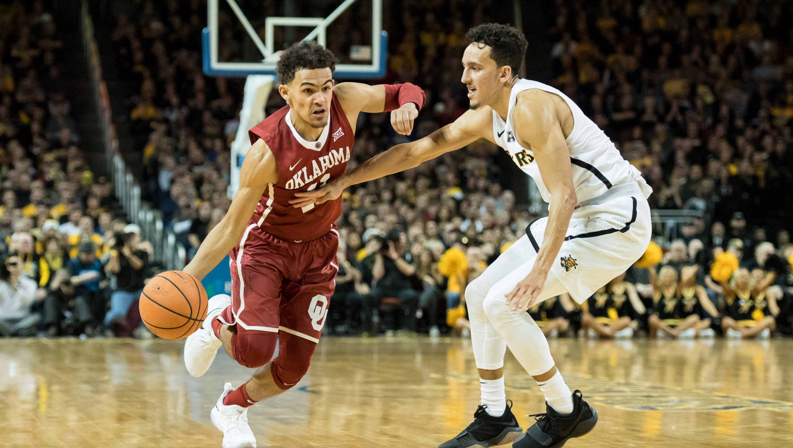 Trae Young 桌布 壁紙 — 一名身穿紅色奧克拉荷馬球衣的籃球運動員在激烈的比賽中突破一名身穿白色球衣的防守球員。