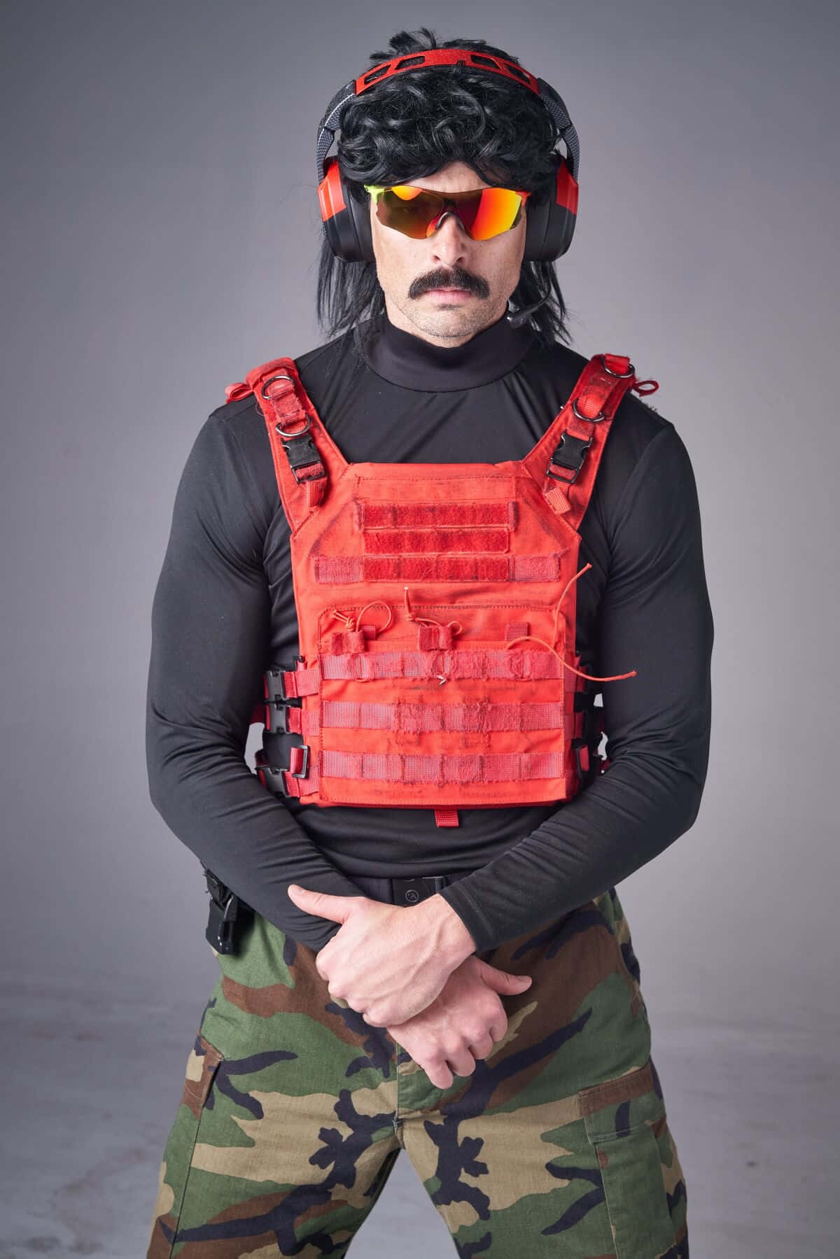 Dr Disrespect 桌布 壁紙 — 一名身穿紅色戰術背心和迷彩褲的男子，戴著黑色假髮和反光太陽眼鏡，在灰色攝影棚背景前擺姿勢。
