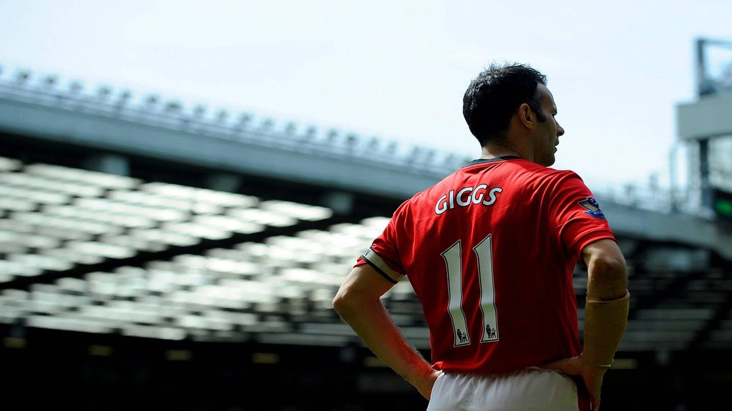 Ryan Giggs 桌布 壁紙 — 一名身穿紅色曼聯球衣、印有「GIGGS」及11號的足球員，雙手叉腰，在明亮的日光下凝視空蕩蕩的球場座位。