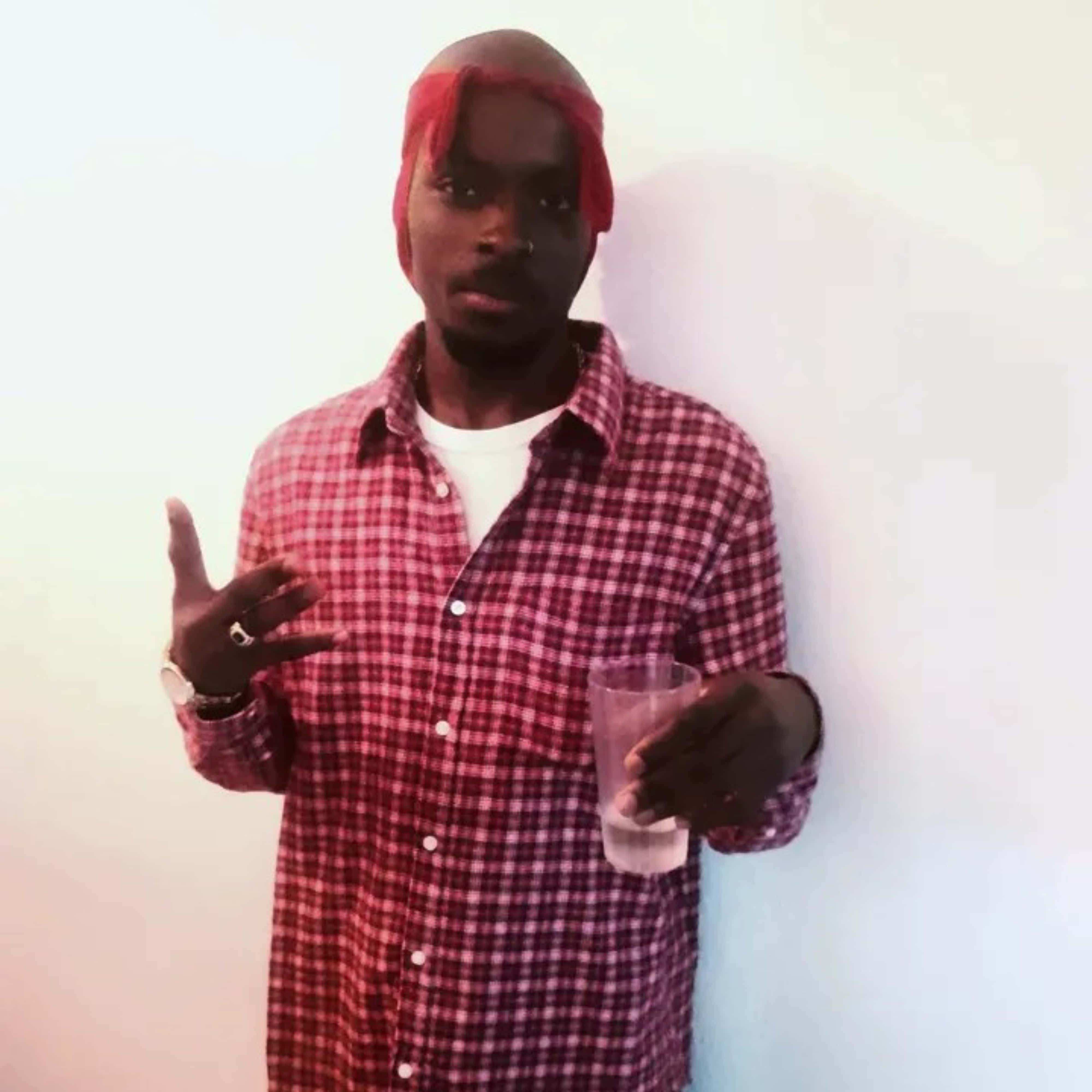 Rap 桌布 壁紙 — 一名身穿紅色格子襯衫和紅色頭巾的男子，手持一杯水，另一隻手在普通牆壁前做手勢。