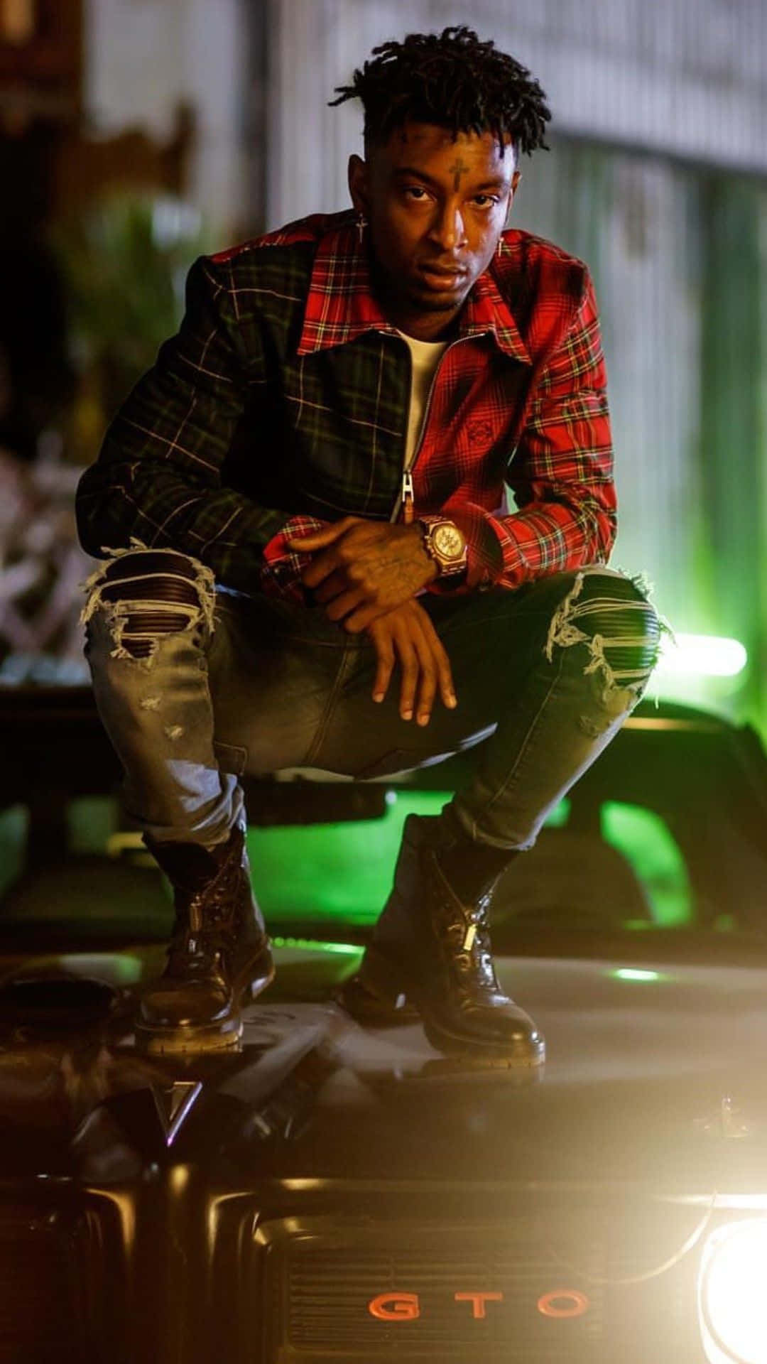 21 Savage 桌布 壁紙 — 一名身穿紅色格紋外套和破洞牛仔褲的年輕男子，在霓虹綠燈光下自信地蹲在一輛黑色GTO汽車上。