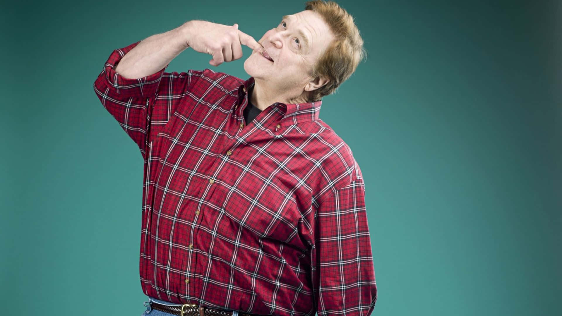John Goodman 桌布 壁紙 — 一名身穿紅色格紋襯衫的男子，在青綠色工作室背景前，帶著俏皮表情指著自己的鼻子。