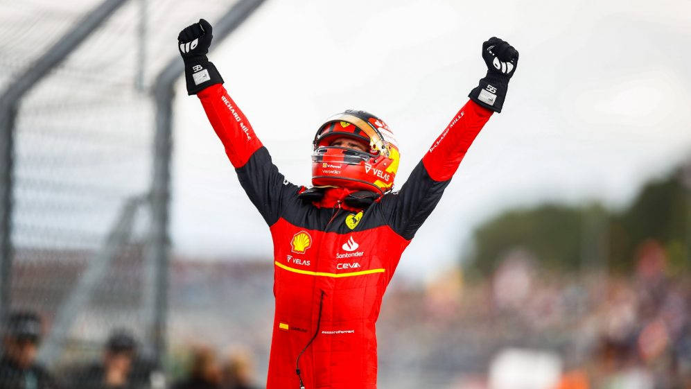 Victory 桌布 壁紙 — 一名身穿紅色法拉利賽車服的F1車手，戴著頭盔，在模糊的賽道背景下高舉雙拳慶祝勝利。
