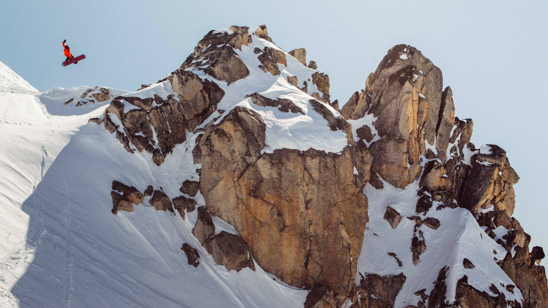Snowboarding 桌布 壁紙 — 一名身穿紅色滑雪服的滑雪板運動員在晴朗的藍天下，飛越崎嶇積雪的山峰。