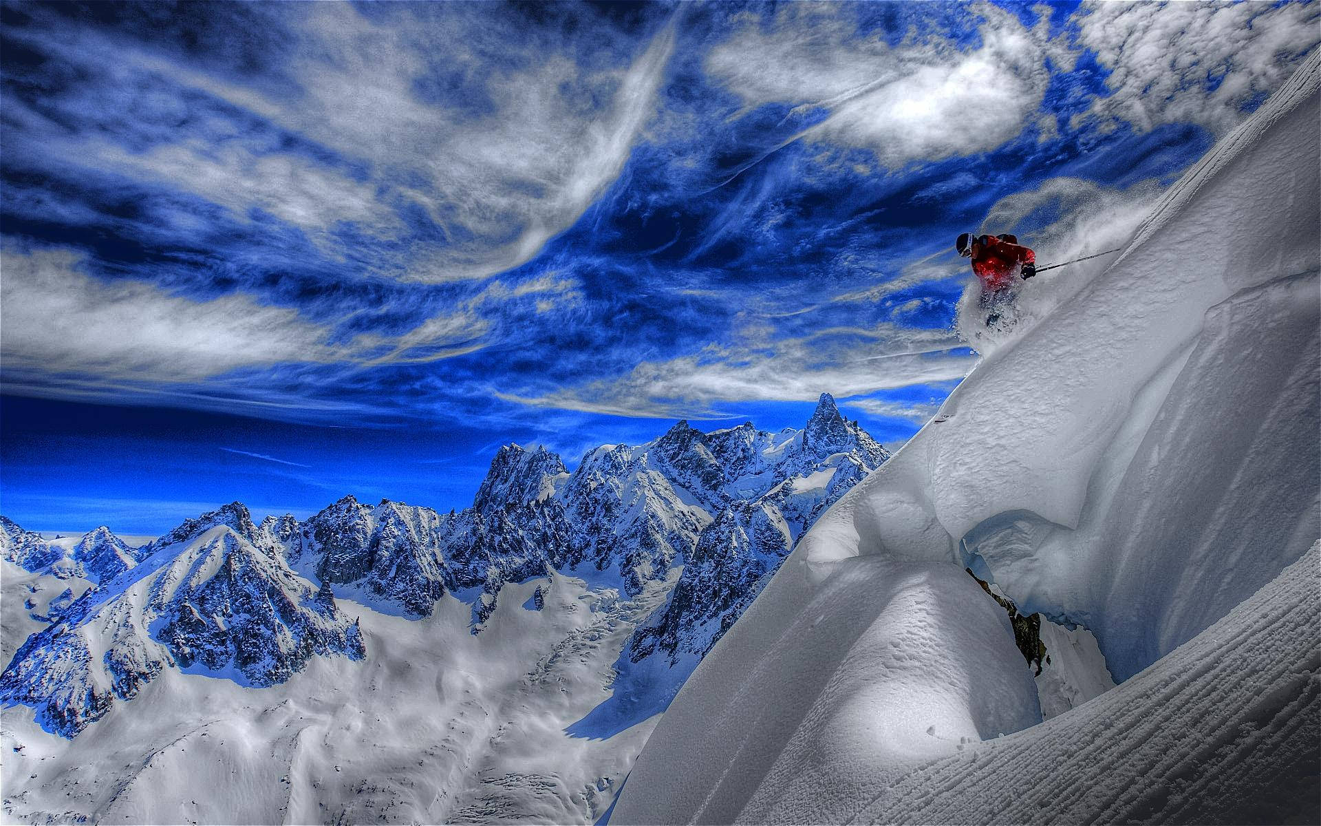 雪山 桌布 壁紙 — 一名身穿紅色滑雪服的滑雪者在陡峭的山脊上穿過深粉雪，背景是戲劇性的藍天和縷縷白雲。