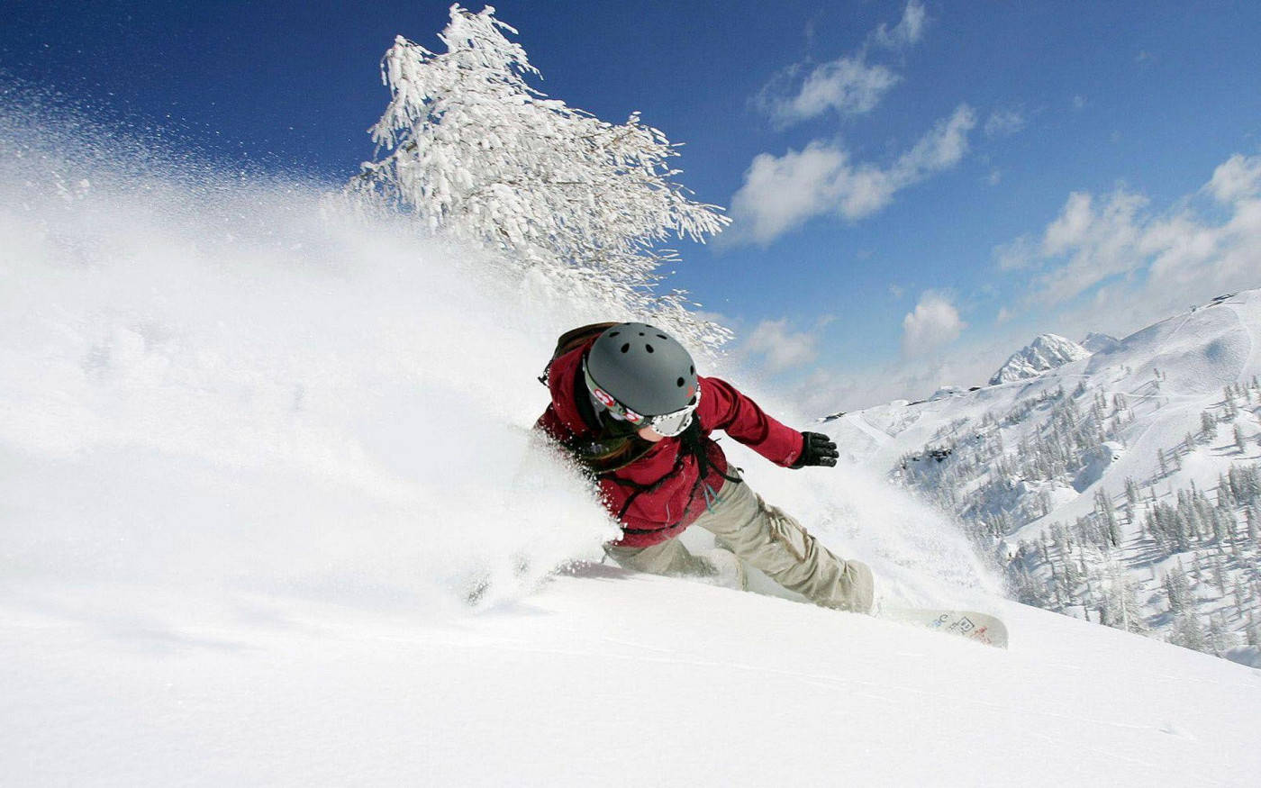  壁紙 — 一名身穿紅色滑雪板的滑雪者在陽光照耀的山坡上，穿過新鮮的粉雪，周圍是白雪覆蓋的樹木和藍天。