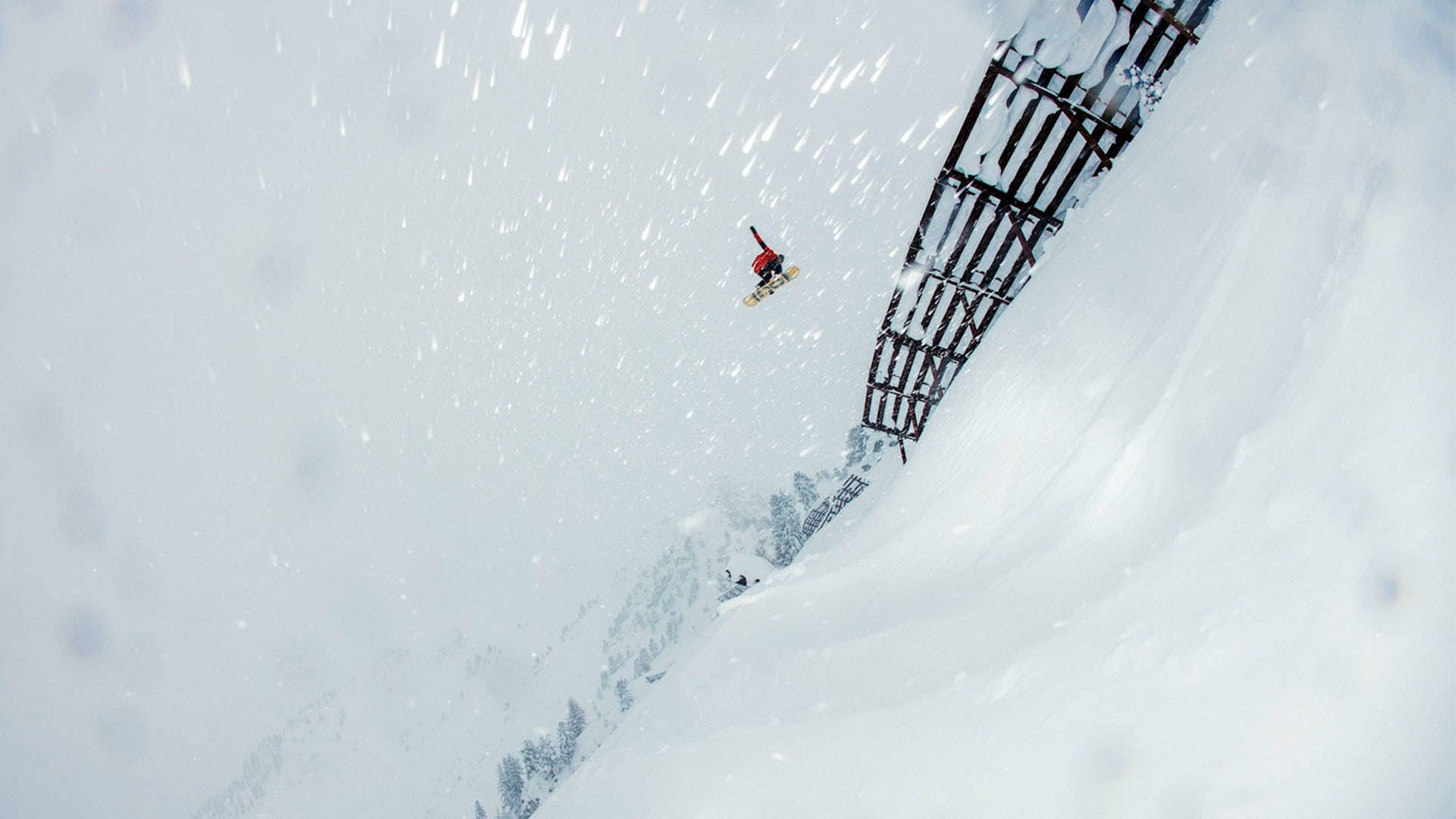 Snowboarding 桌布 壁紙 — 一名身穿紅色滑雪板運動員在陡峭的雪山斜坡上，在金屬欄杆附近飛過飄落的雪花。