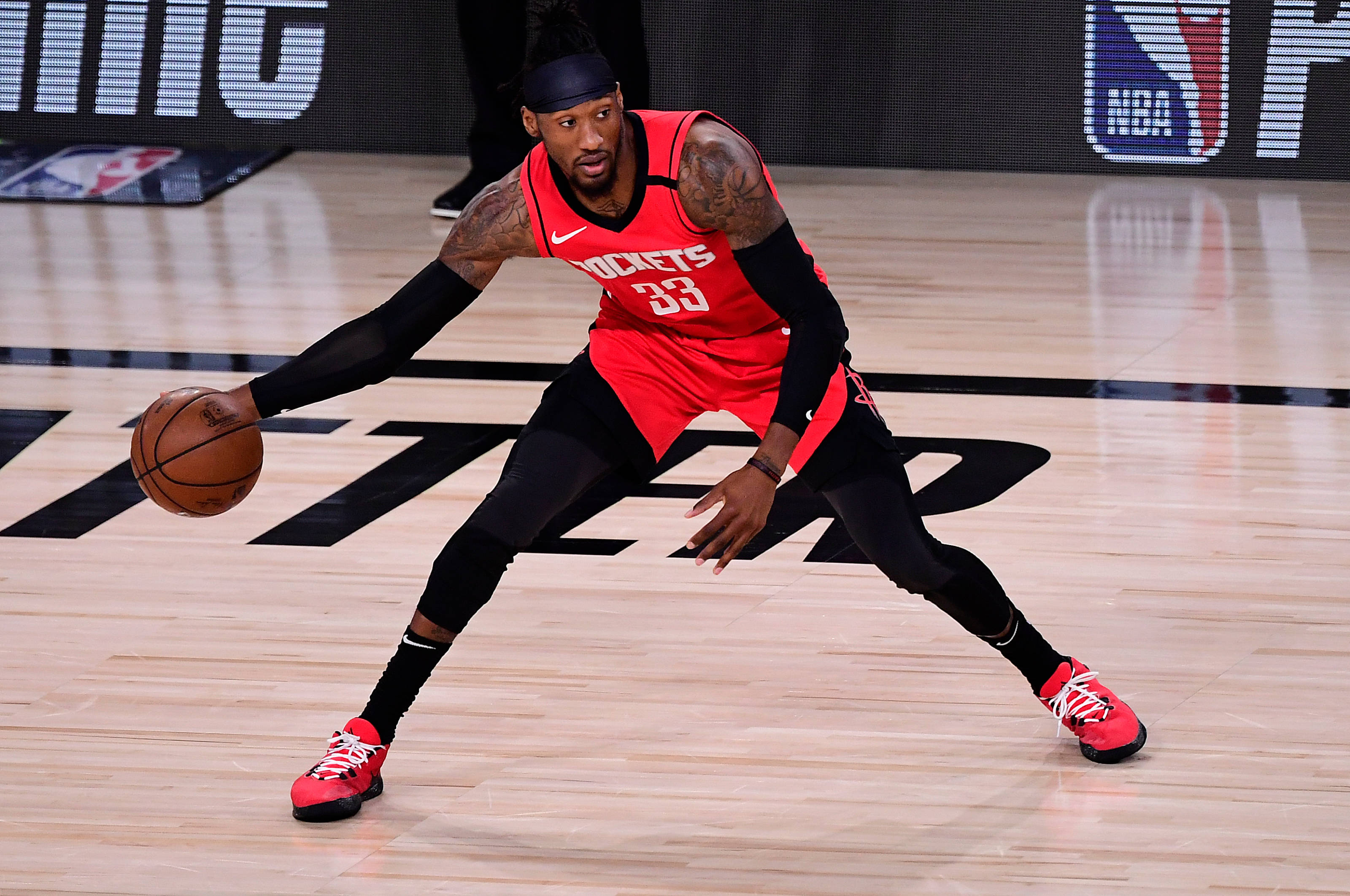 robert covington 桌布 壁紙 — 一名身穿紅色火箭隊球衣的籃球運動員在明亮的場館燈光下，專注地在拋光木地板上運球。
