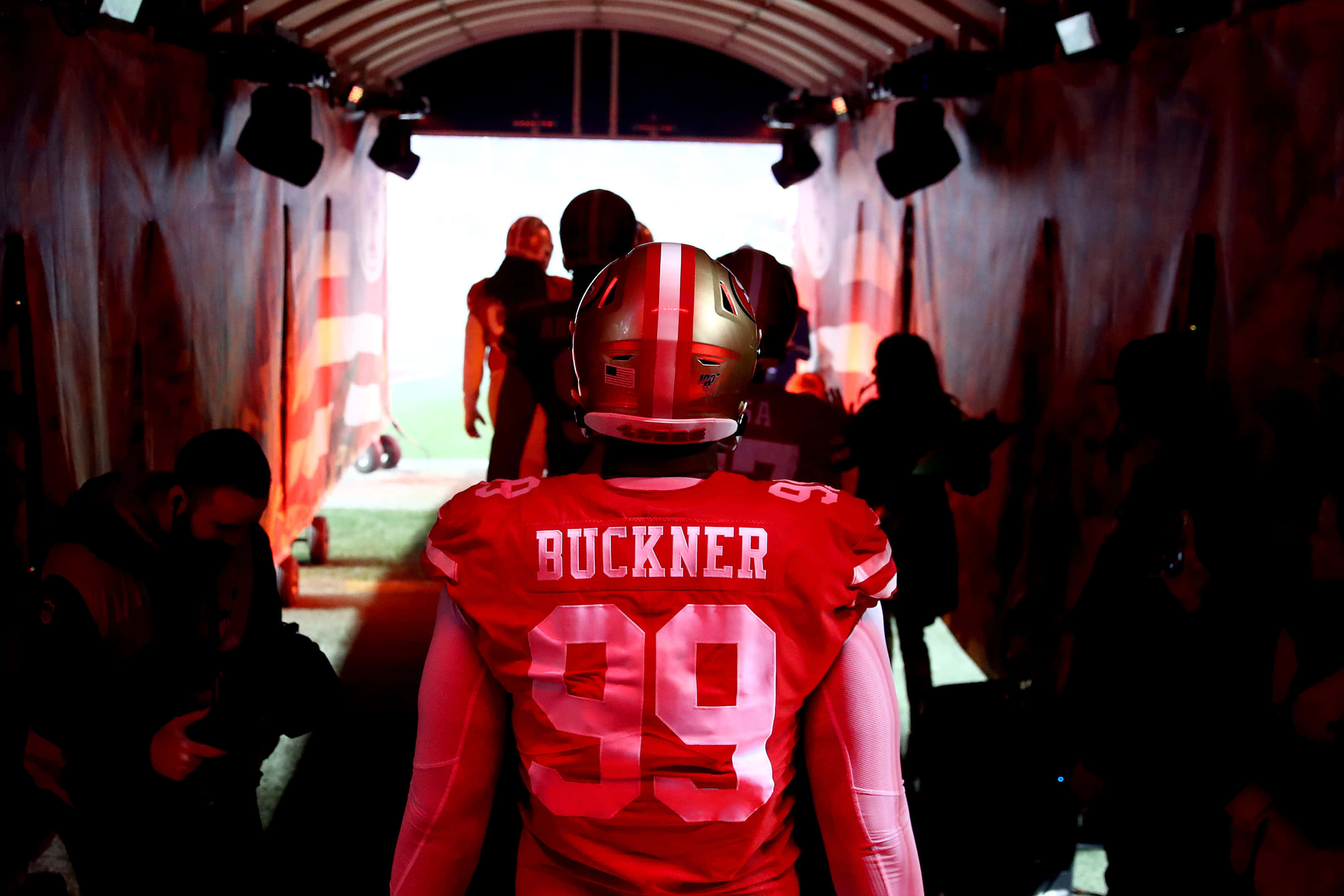 Football Players 桌布 壁紙 — 一名身穿紅色球衣、名字為Buckner、號碼99的美式足球員，在戲劇性的紅光下穿過隧道走向球場。