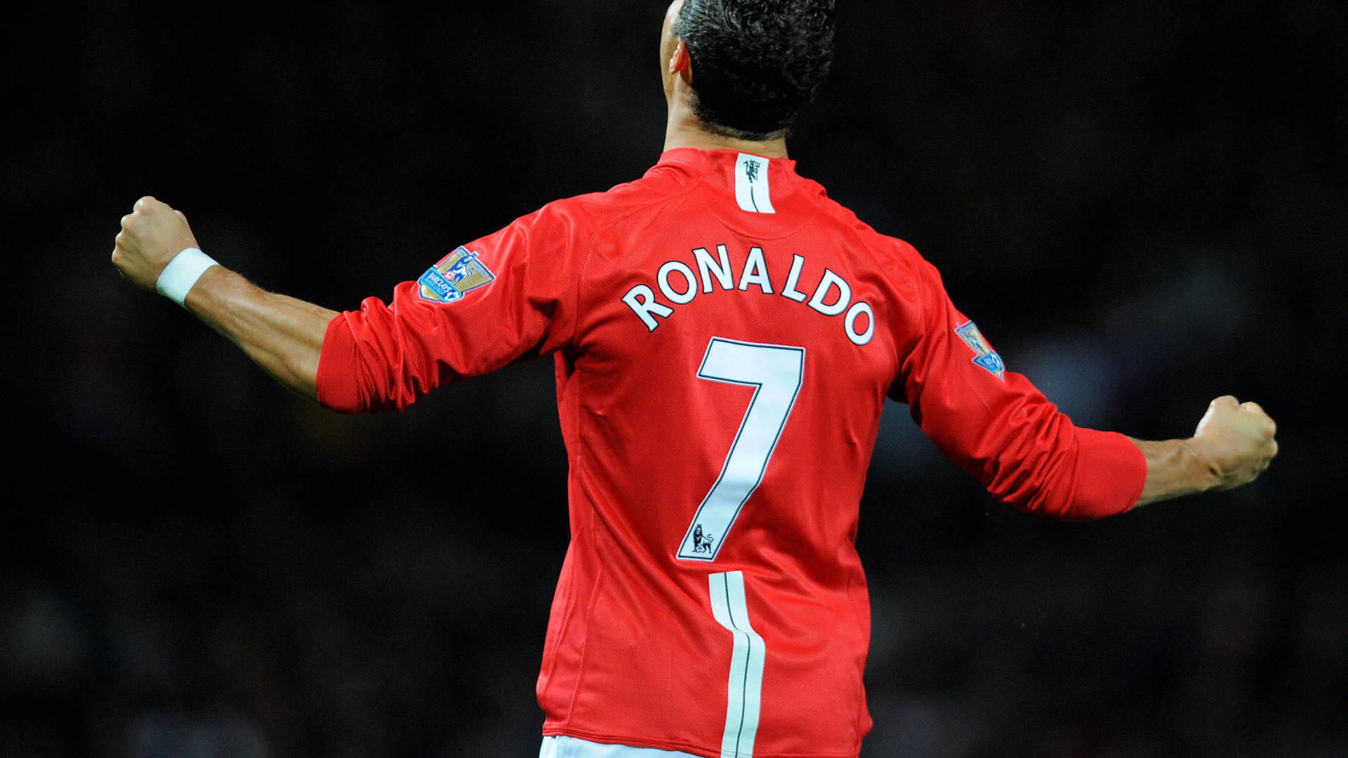 克里斯蒂亞諾·羅納度 桌布 壁紙 — 一名身穿紅色球衣、背後印有「RONALDO」和7號的足球運動員在黑暗的球場背景下張開雙臂慶祝。