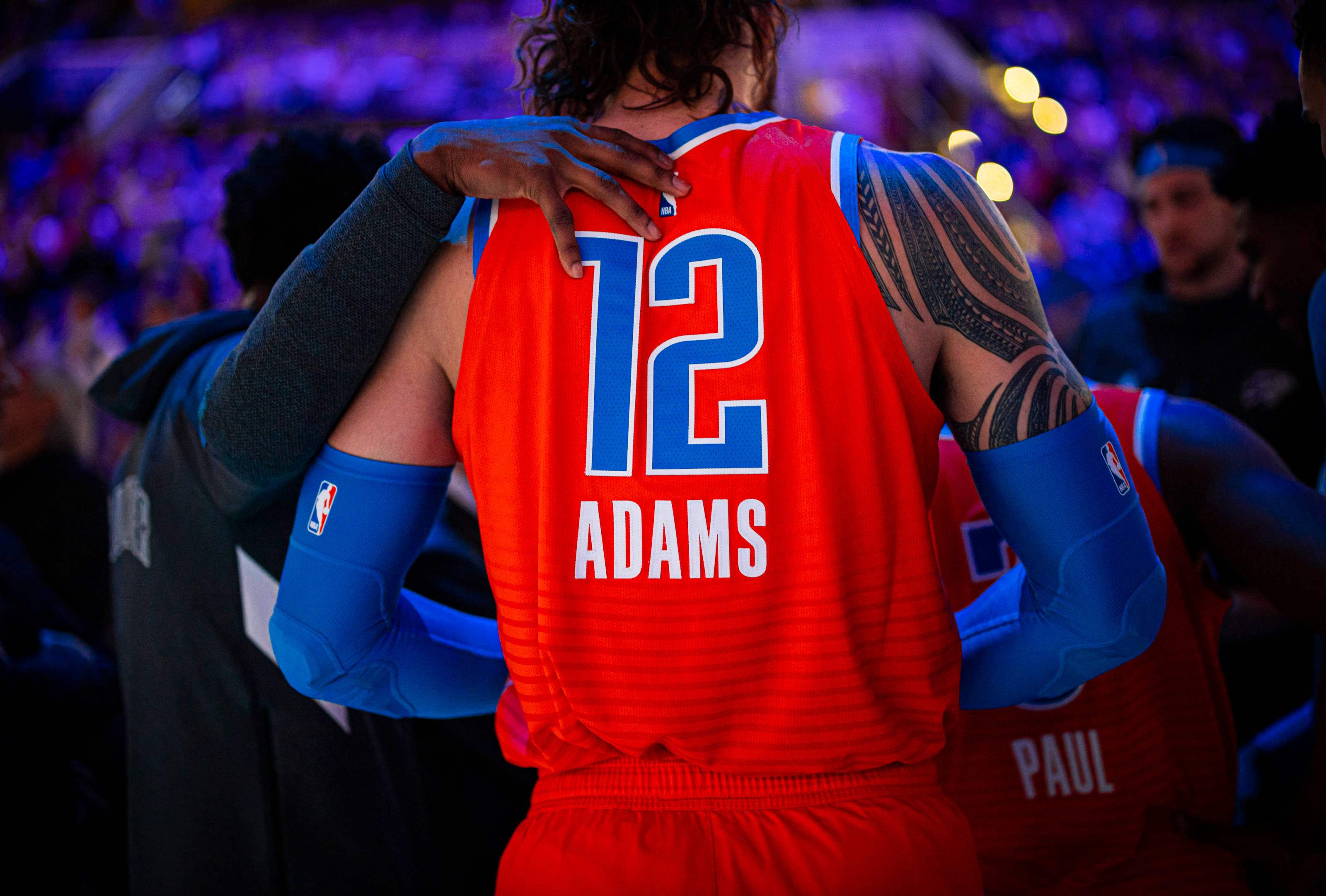 Steven Adams 桌布 壁紙 — 一名身穿紅色球衣、背號12、名字Adams的籃球運動員，在昏暗的場館中被隊友從背後擁抱。