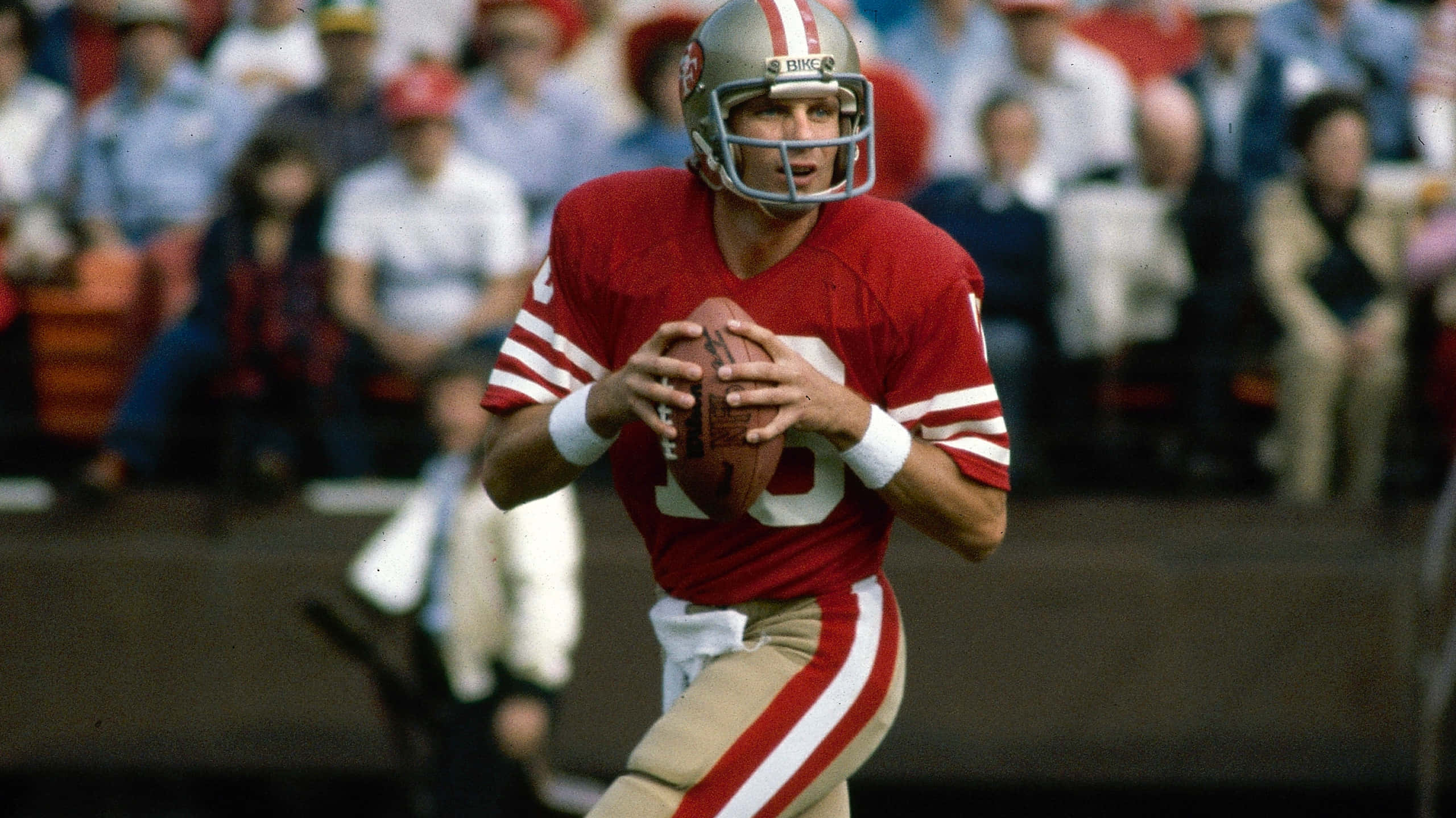 Joe Montana 桌布 壁紙 — 一名身穿紅色球衣和頭盔的美式足球四分衛在觀眾背景下準備在比賽中擲球。