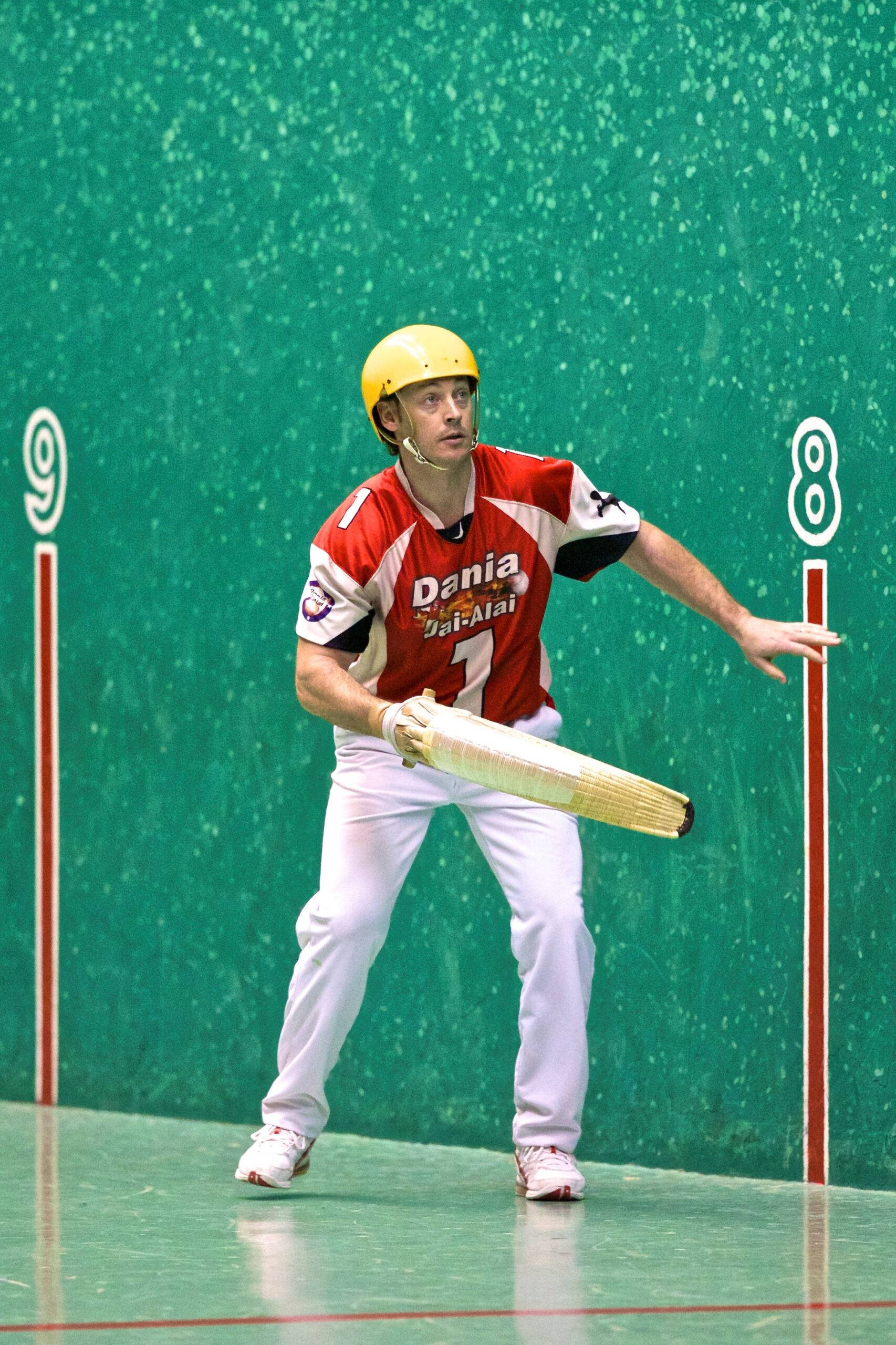 Athletic 桌布 壁紙 — 一名身穿紅色球衣和黃色頭盔的男性壁球選手，正準備用長球拍擊球，對著一面標有數字的綠色紋理牆壁。