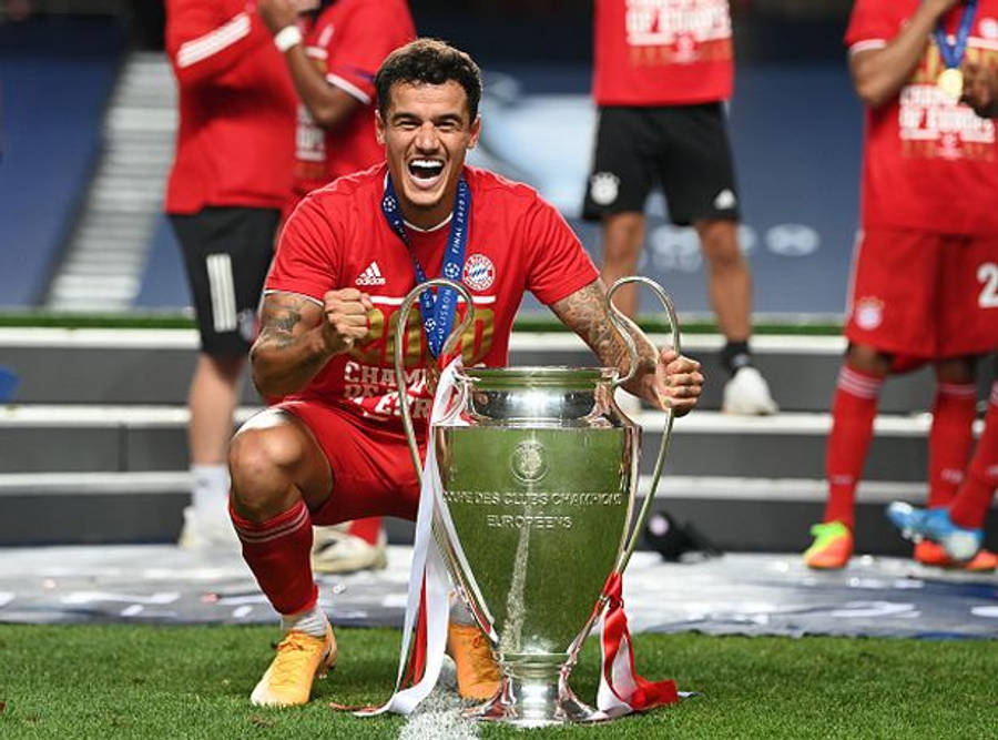 Philippe Coutinho 桌布 壁紙 — 一名身穿紅色球衣的喜悅足球員跪在草地上，手持閃閃發光的歐冠獎盃，隊友在他身後慶祝。