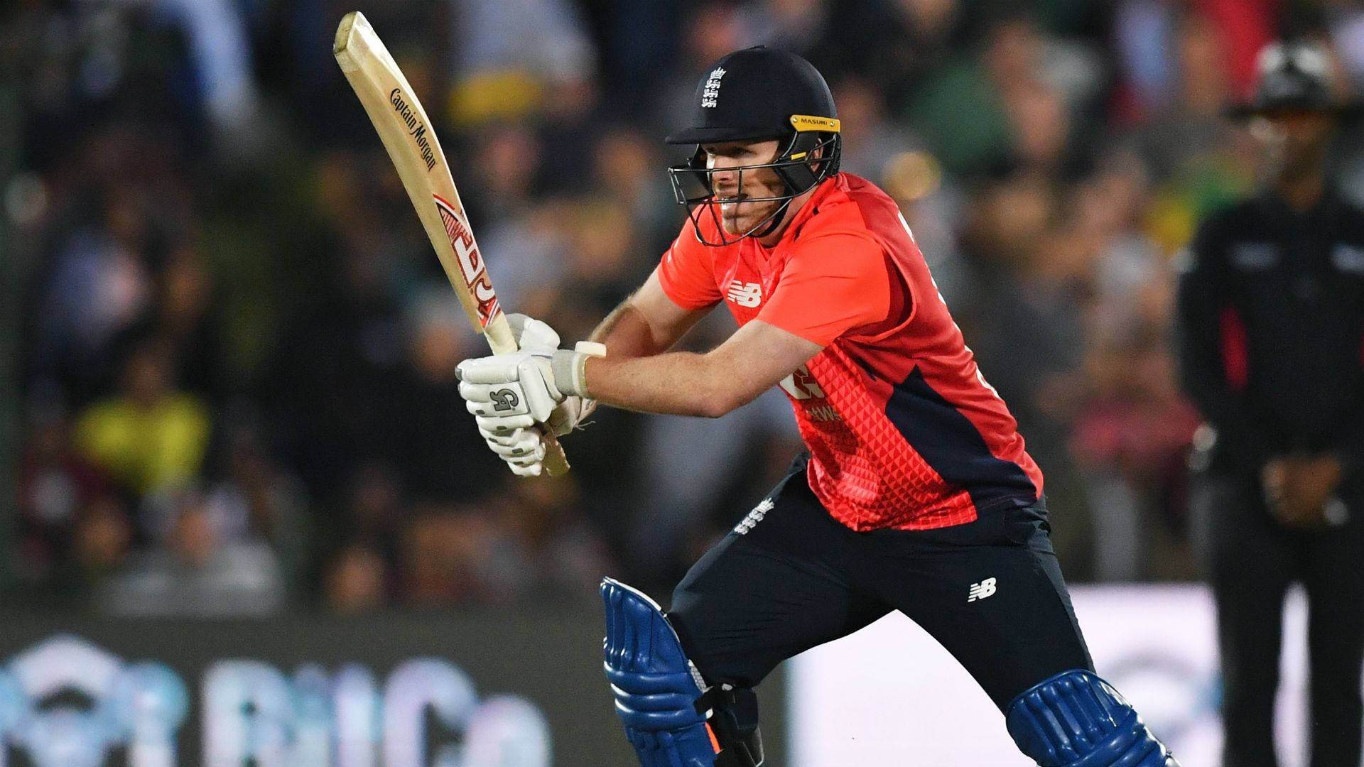 eoin morgan 桌布 壁紙 — 一名身穿紅色球衣的板球擊球手在夜間比賽中，在體育場燈光下強力揮棒。