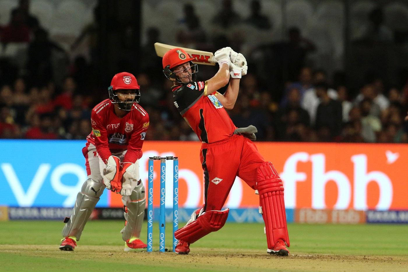 royal challengers bangalore 桌布 壁紙 — 一名身穿紅色球衣的板球擊球手在夜間比賽中強力揮動球棒，而守門員蹲在門柱後面。