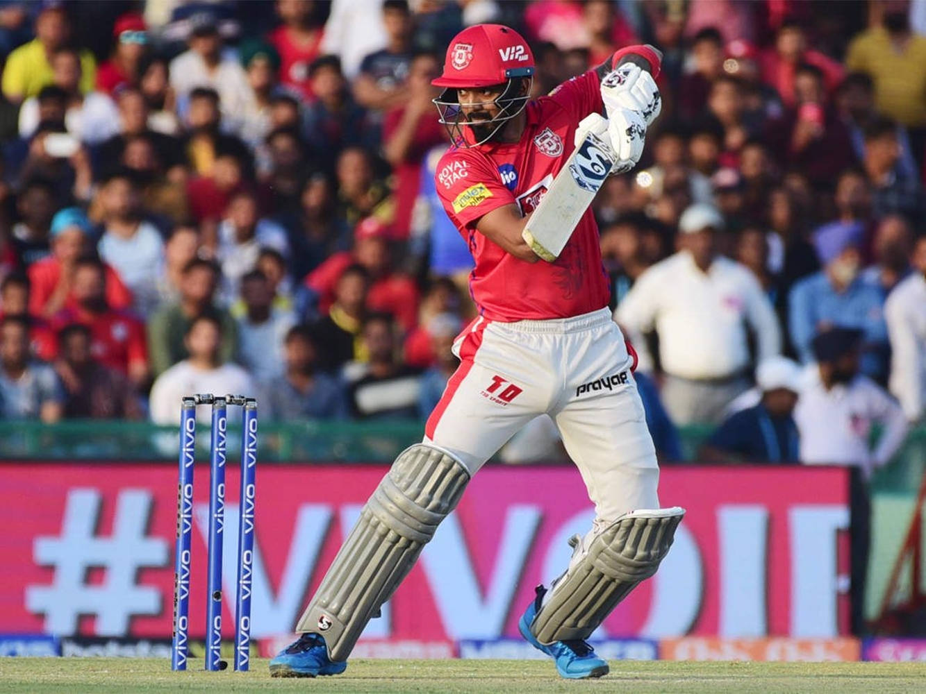 kl rahul 桌布 壁紙 — 一名身穿紅色球衣的板球擊球手在日間比賽中強力揮棒，身後是座無虛席的體育場觀眾。