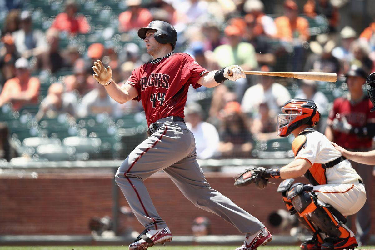Paul Goldschmidt 桌布 壁紙 — 一名身穿紅色球衣的棒球運動員在揮棒中途揮動球棒，而捕手則在陽光明媚的球場上蹲在他身後。