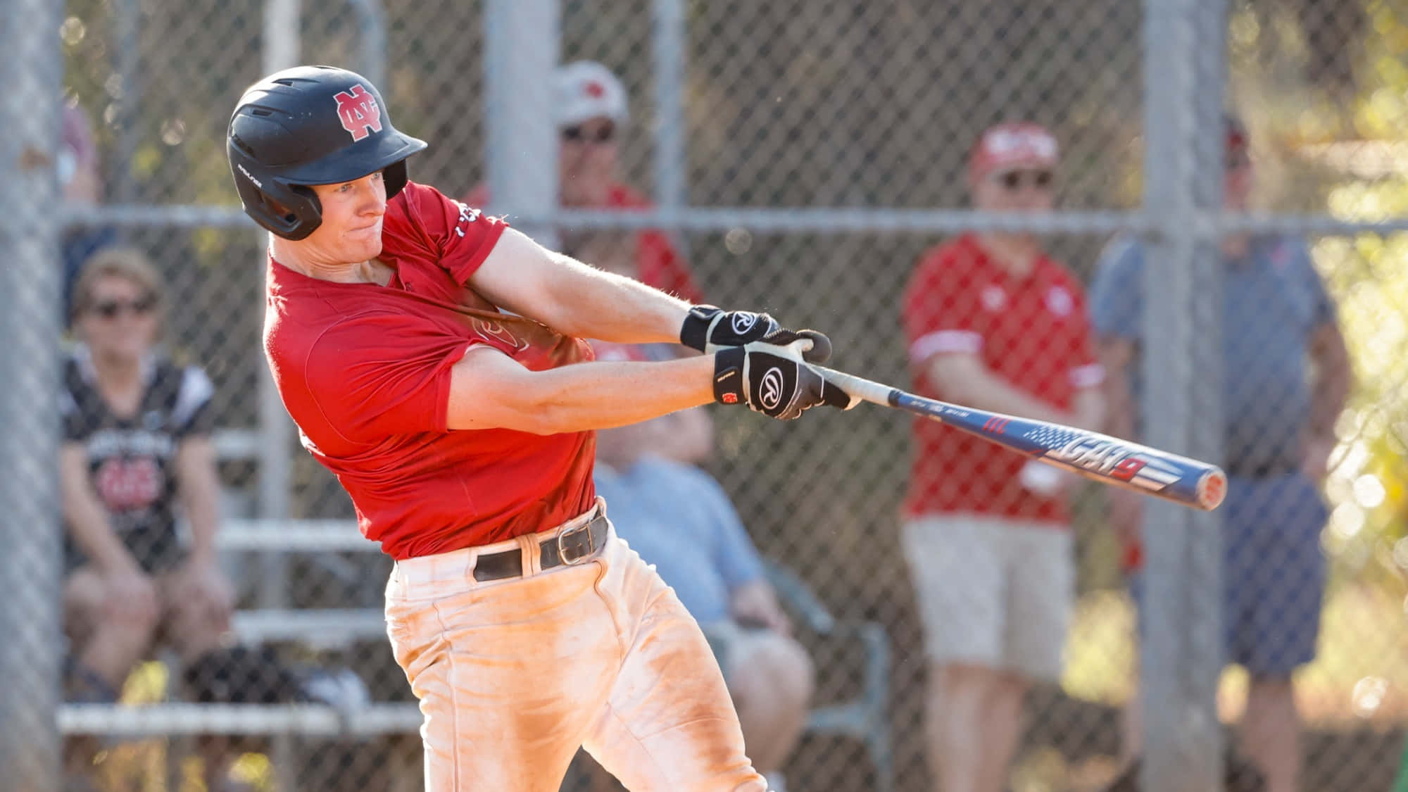 College Baseball 桌布 壁紙 — 一名身穿紅色球衣的棒球選手在日間比賽中，專注地揮動球棒，鐵絲網圍欄後有觀眾觀看。