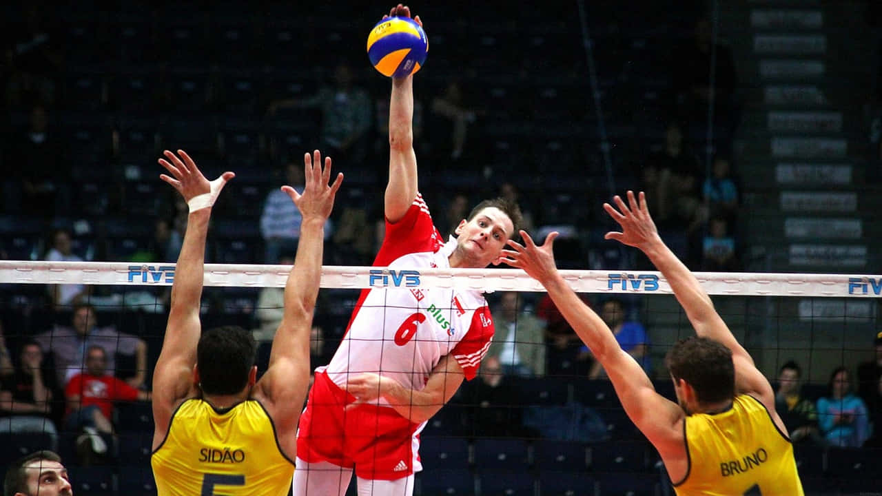 Volleyball 桌布 壁紙 — 一名身穿紅色球衣的男排球員將球扣過網，兩名身穿黃色球衣的對手試圖攔網。