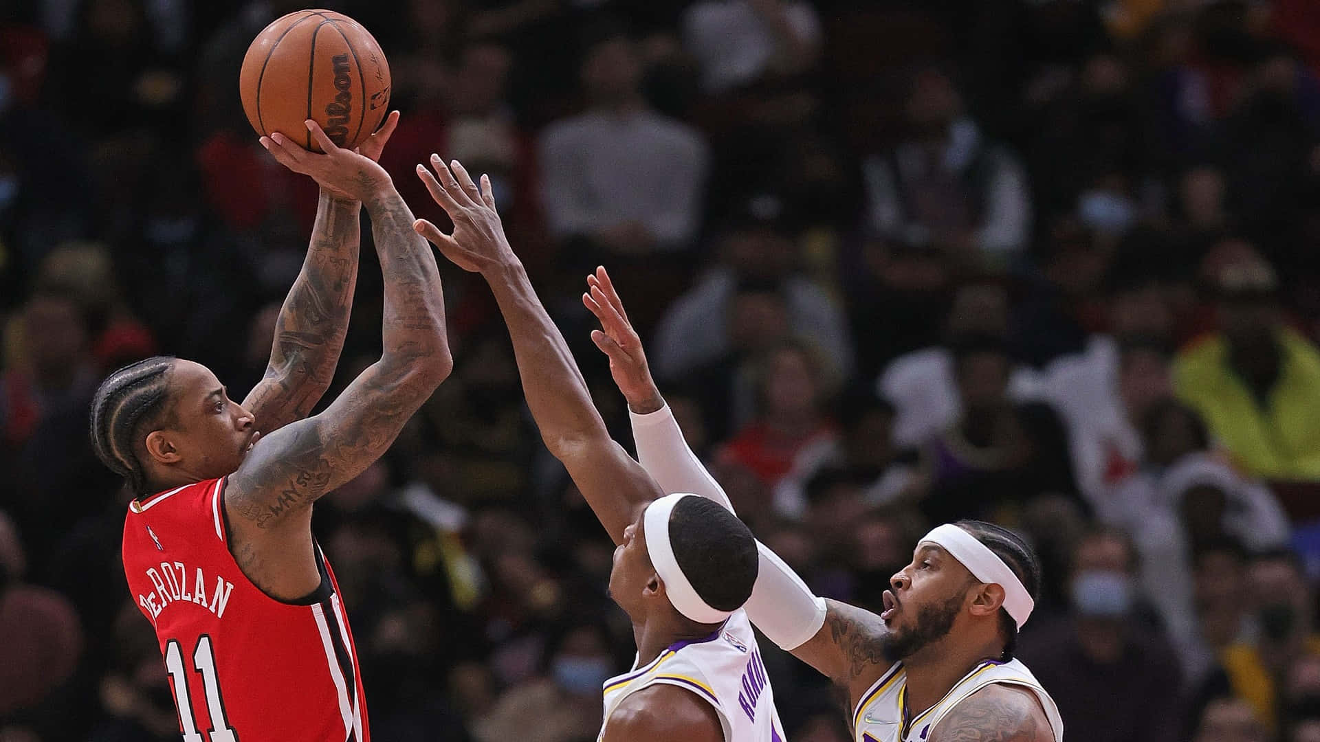 NBA 桌布 壁紙 — 一名身穿紅色球衣的籃球運動員在明亮的燈光下，於擠滿觀眾的場館內越過兩名防守球員投籃。