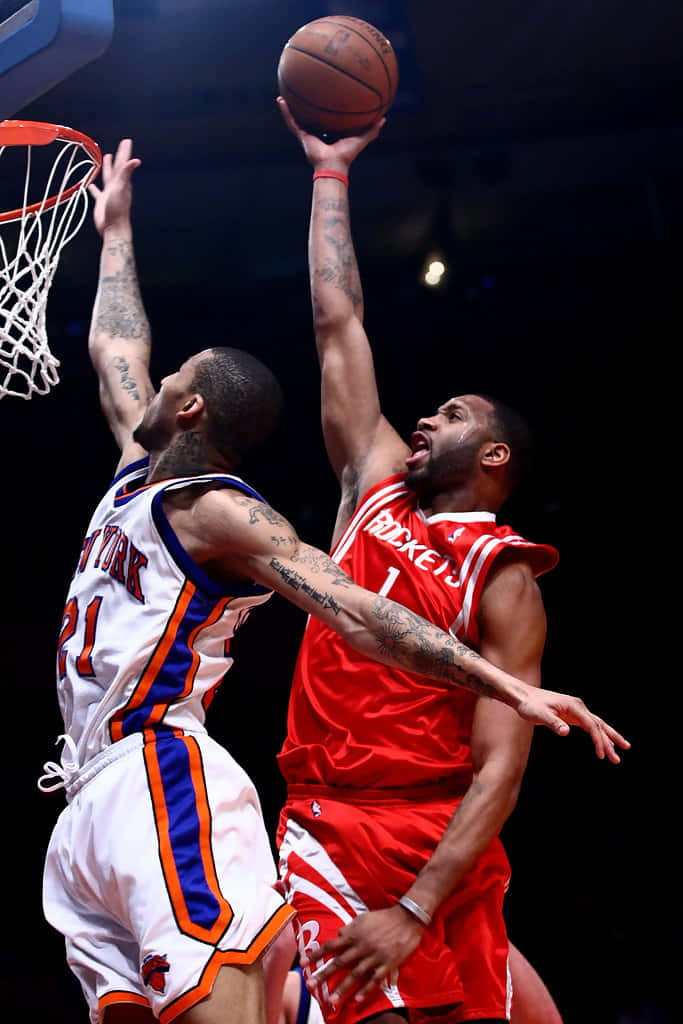 Tracy Mcgrady 桌布 壁紙 — 一名身穿紅色球衣的籃球運動員跳起灌籃，而一名身穿白色球衣的防守球員在場館燈光下試圖封阻。