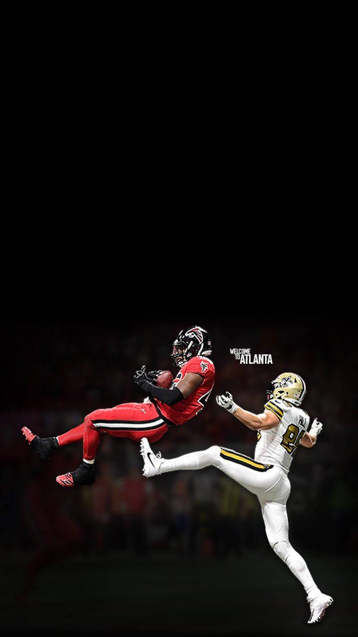 Football Players 桌布 壁紙 — 一名身穿紅色球衣的美式足球員在半空中跳起接球，而一名身穿白色球衣的防守球員在球場燈光下撲向他試圖阻止。