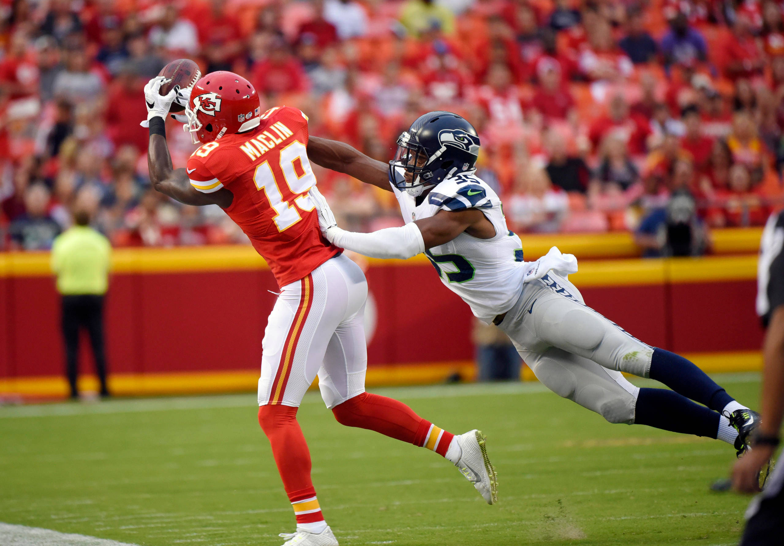 Kansas City Chiefs Cool 桌布 壁紙 — 一名身穿紅色球衣的美式足球員接住傳球，而一名身穿白色球衣的防守球員在陽光普照的球場上飛身擒抱他。