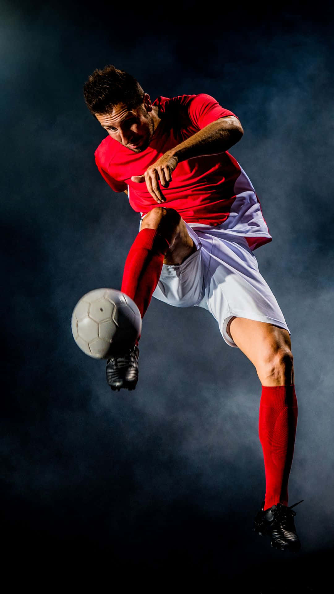 Soccer iPhone 桌布 壁紙 — 一名身穿紅色球衣的足球員在半空中跳起，在煙霧瀰漫的深色背景前擊球。