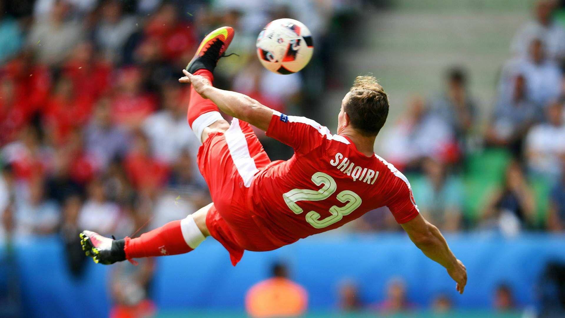足球 桌布 壁紙 — 一名身穿紅色球衣的足球員在日間比賽中，於模糊觀眾前凌空做出倒掛金鉤動作。