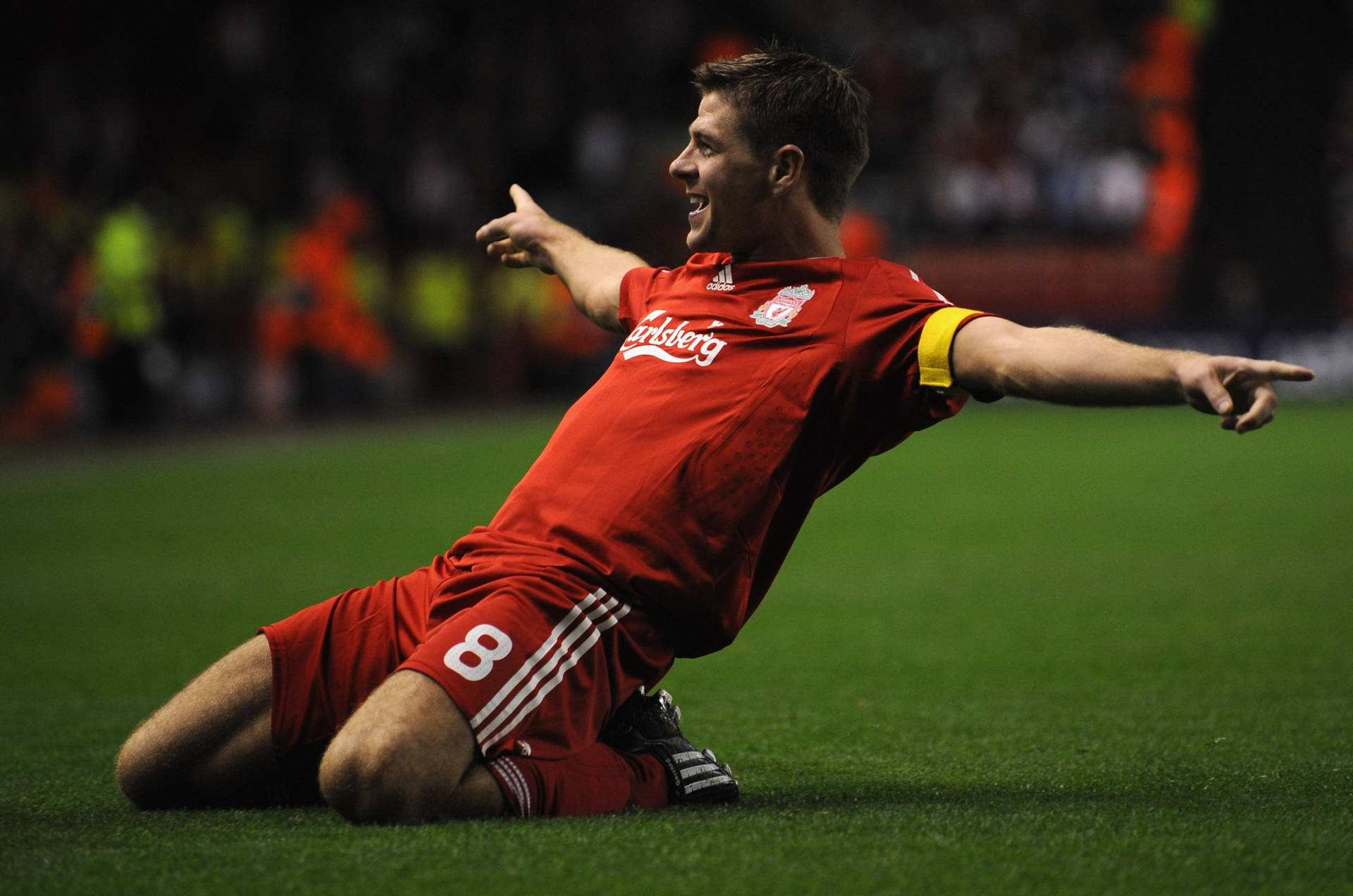 Steven Gerrard 桌布 壁紙 — 一名身穿紅色球衣的足球員在草地滑行，雙臂張開慶祝，球場燈光下。