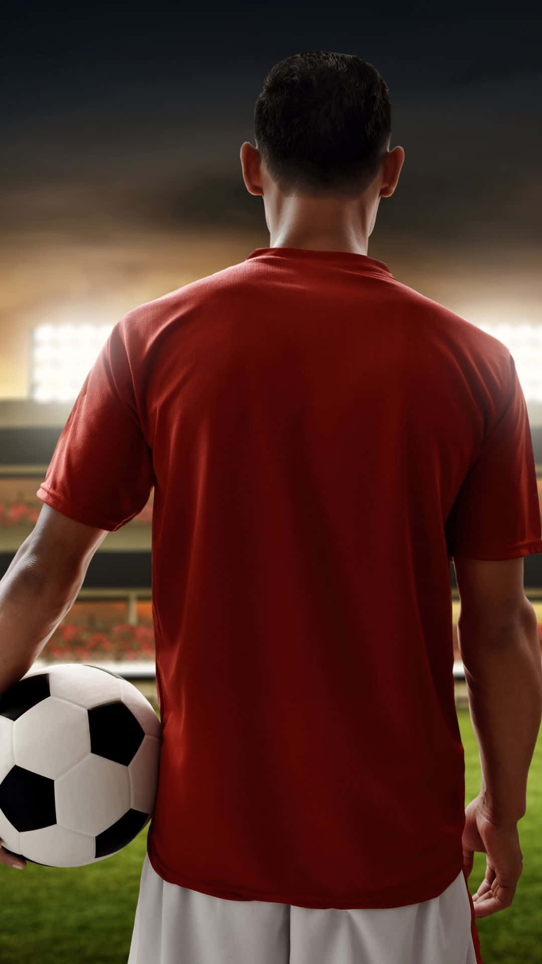 Soccer iPhone 桌布 壁紙 — 一名身穿紅色球衣的足球員站在球場上，在戲劇性的球場燈光下拿著一個足球。