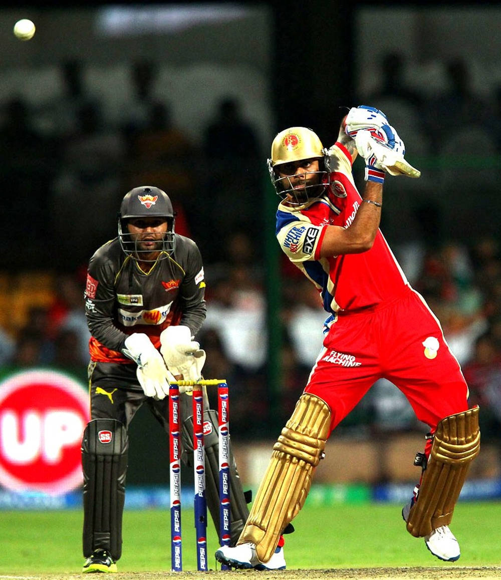 royal challengers bangalore 桌布 壁紙 — 一名身穿紅色的板球擊球手強力揮棒擊球，而一名身穿黑色的守門員在體育場燈光下專注觀看。