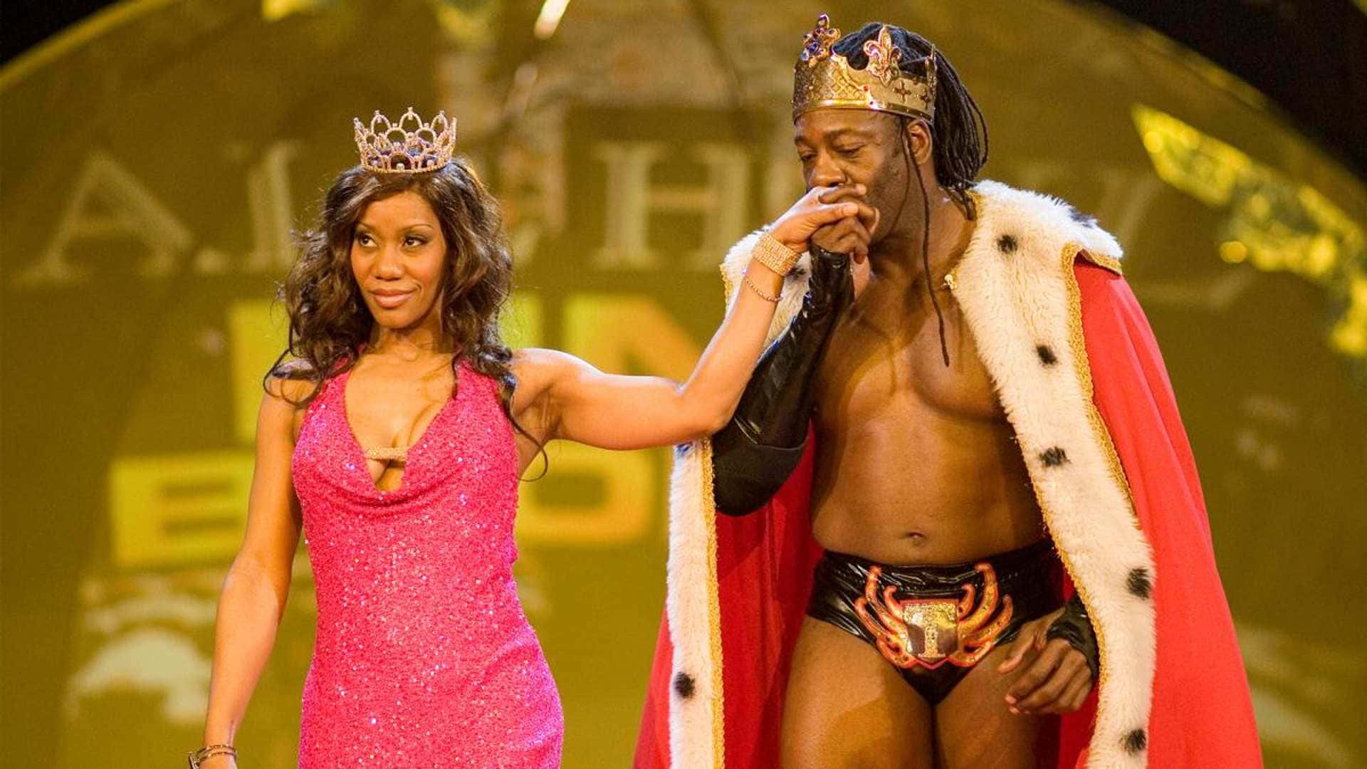 WWE 桌布 壁紙 — 一名身穿紅色皇家斗篷和皇冠的男子站在一名身穿粉紅色亮片連衣裙的女子旁邊，兩人都戴著皇冠站在舞台上。