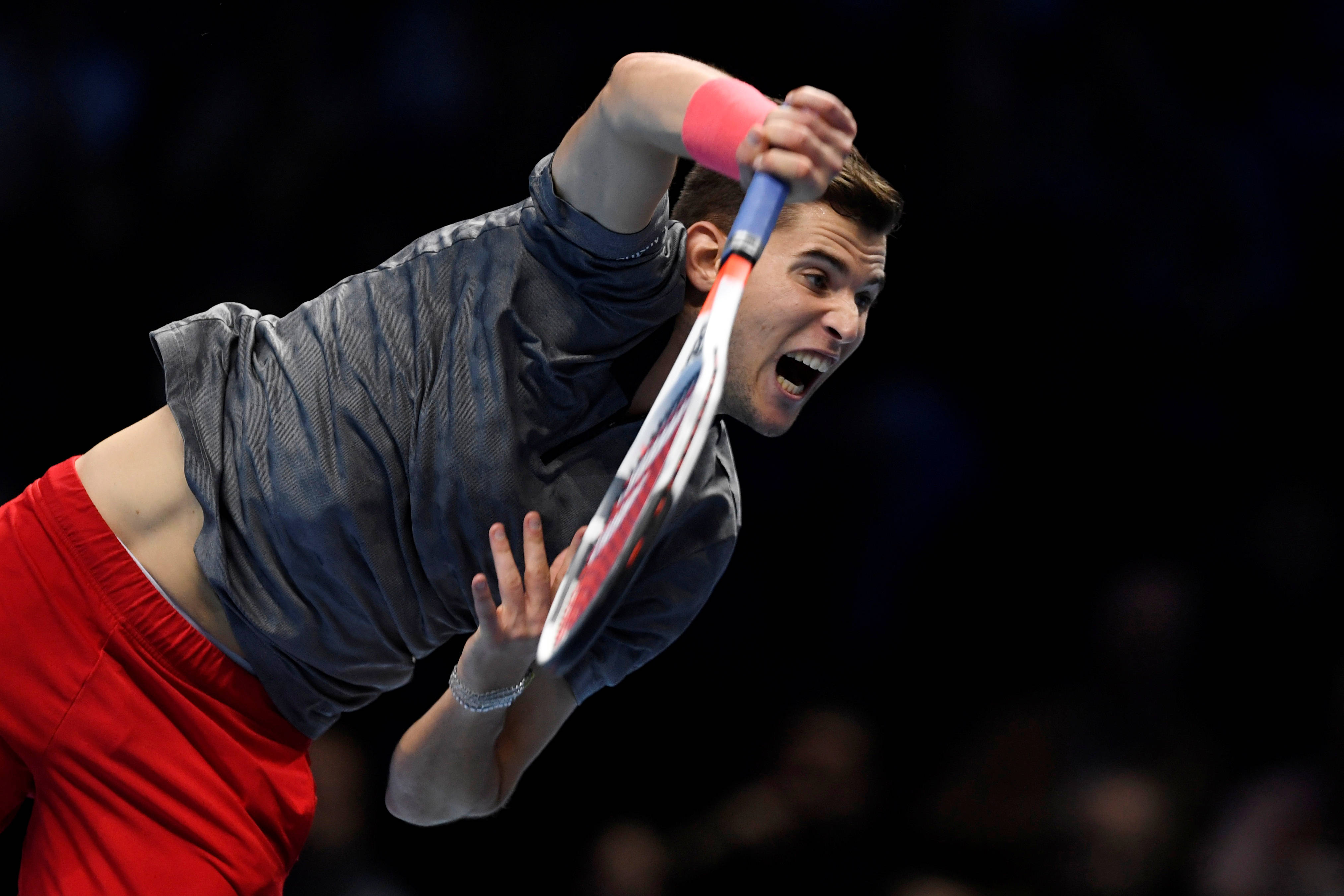 dominic thiem 桌布 壁紙 — 一名身穿紅色短褲和灰色襯衫的男性網球運動員，在黑暗模糊的背景前，以強烈的專注力向前猛衝，揮動他的球拍。