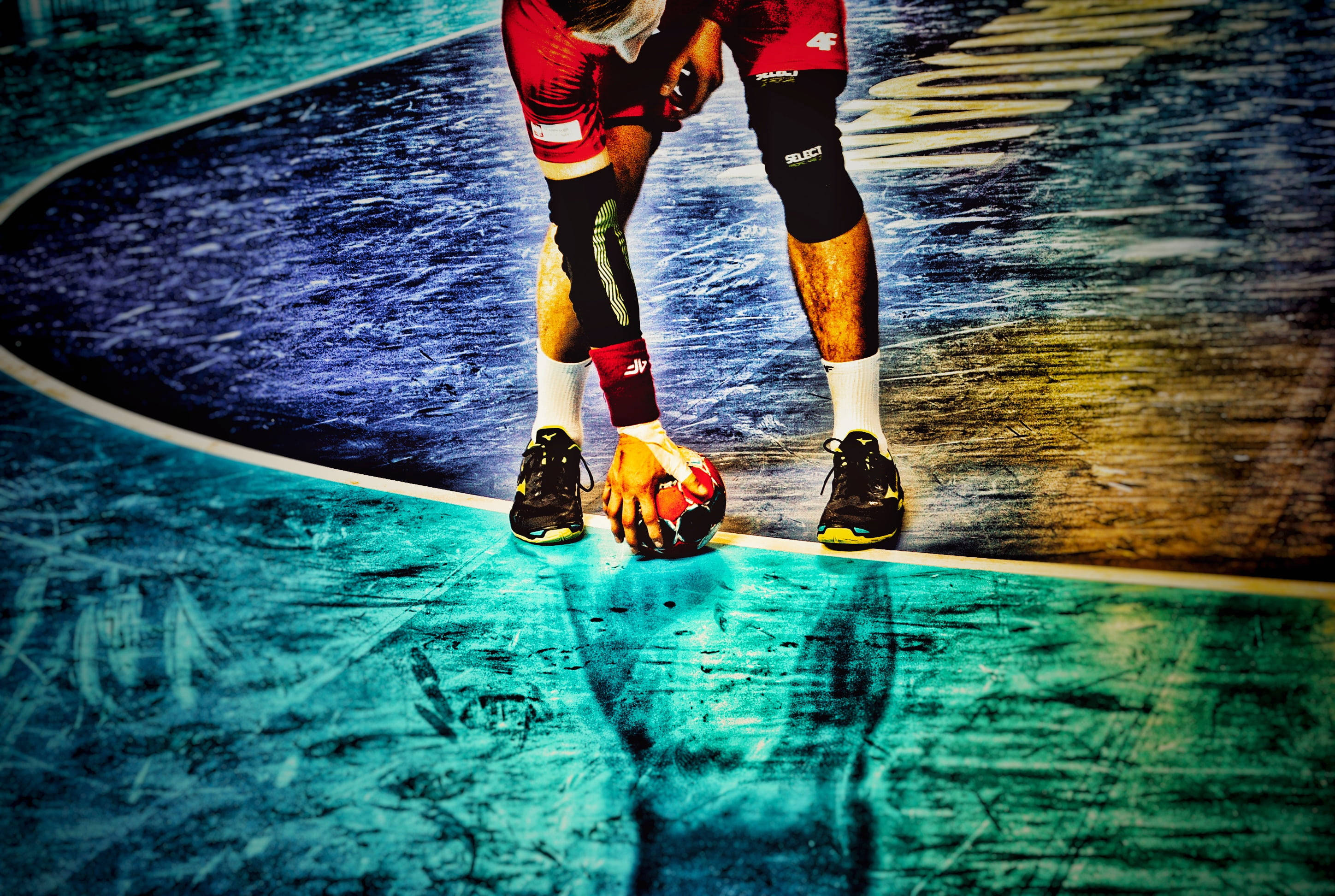 Handball 桌布 壁紙 — 一名身穿紅色短褲和黑色護膝的手球運動員在充滿活力的青綠色和藍色球場上低蹲著拿著球。