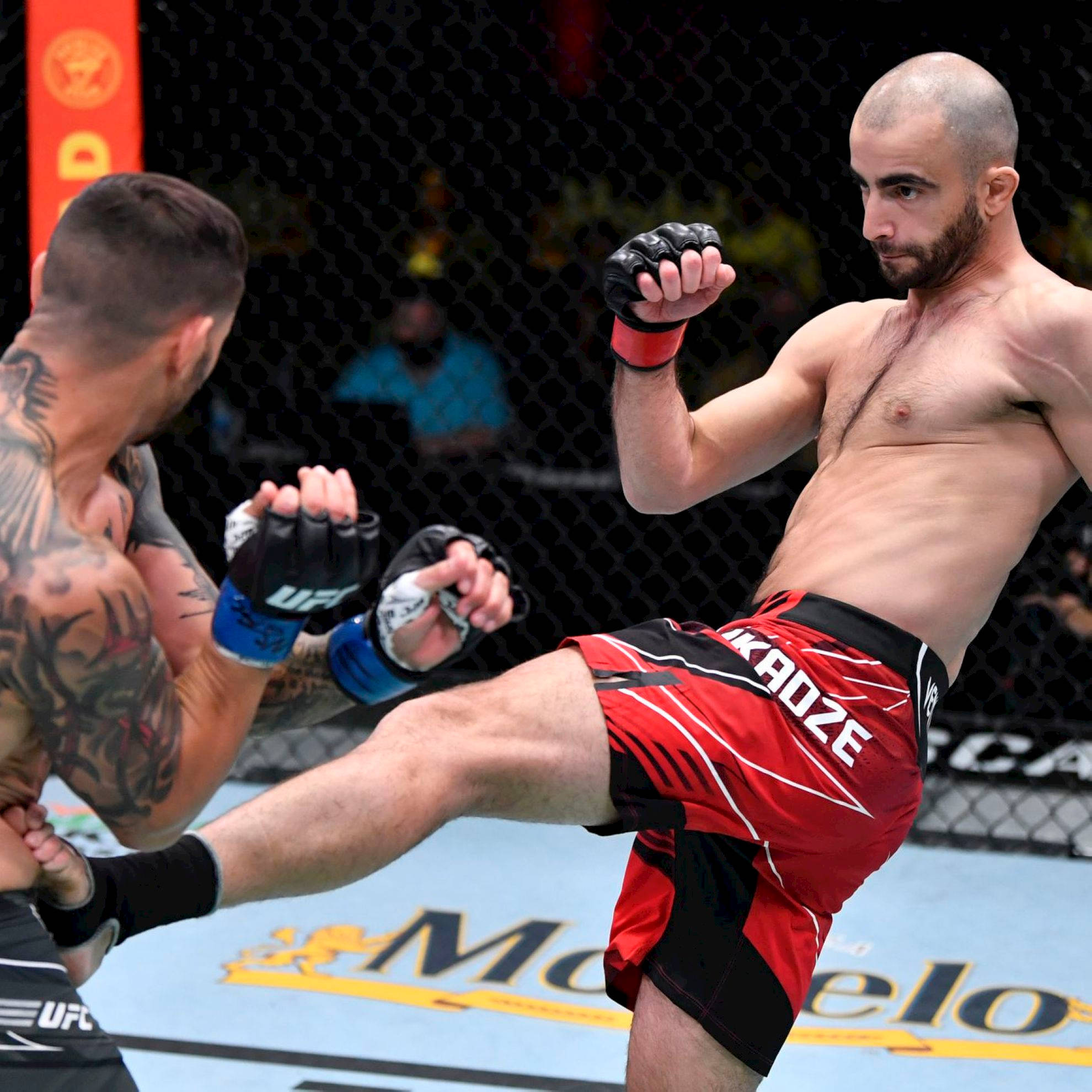 UFC 桌布 壁紙 — 一名身穿紅色短褲的拳手在明亮的場館燈光下，於UFC八角籠內向對手踢出一記高踢。