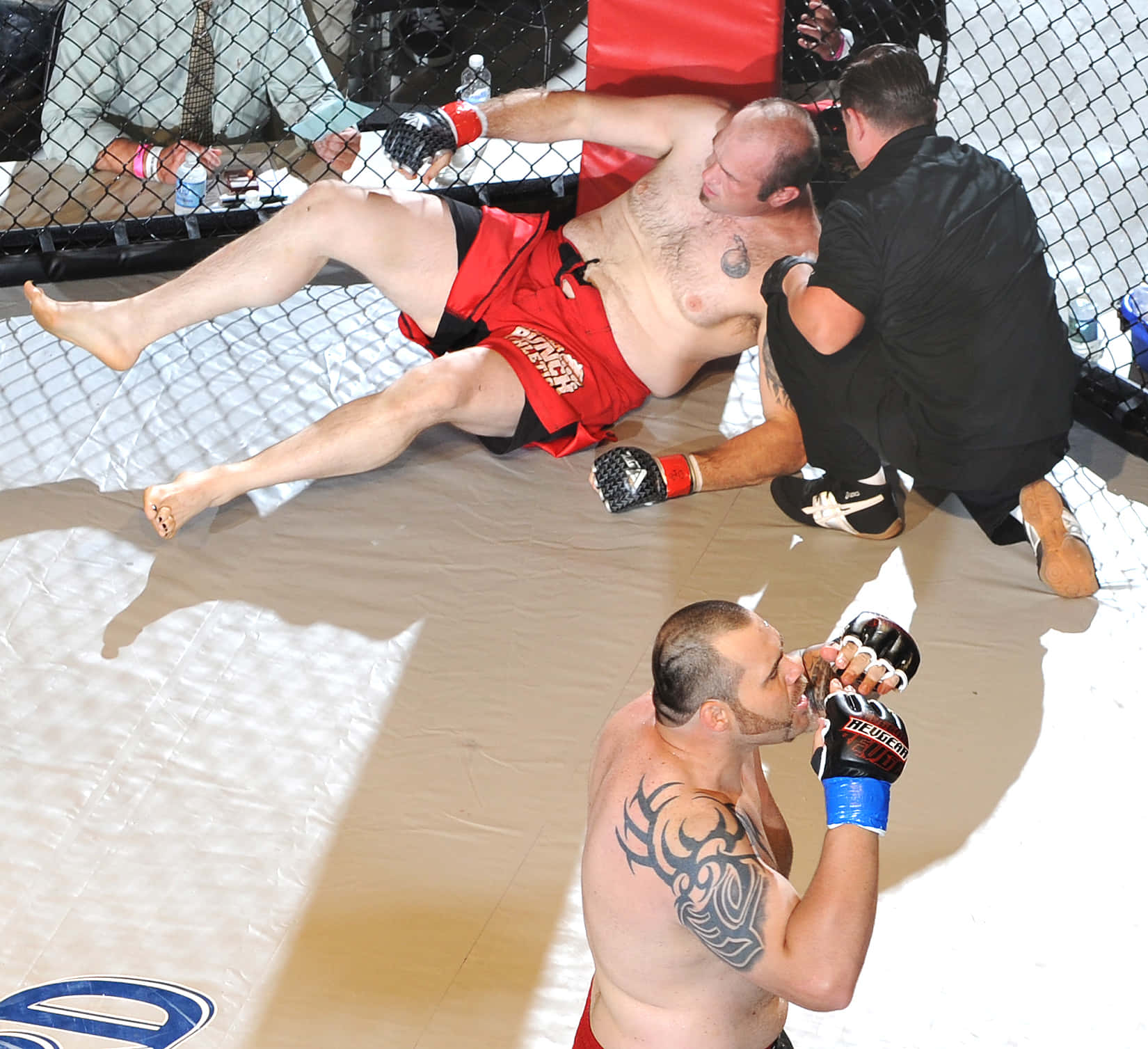 MMA 桌布 壁紙 — 一名身穿紅色短褲的拳手被打倒在墊子上，裁判正在檢查他，另一名拳手站在附近。