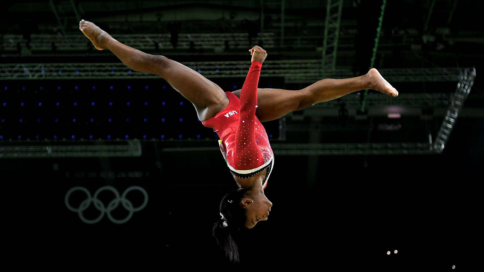 Simone Biles 桌布 壁紙 — 一名身穿紅色緊身衣的女子體操運動員在明亮的場館燈光下，於平衡木上做出完美的倒立動作。