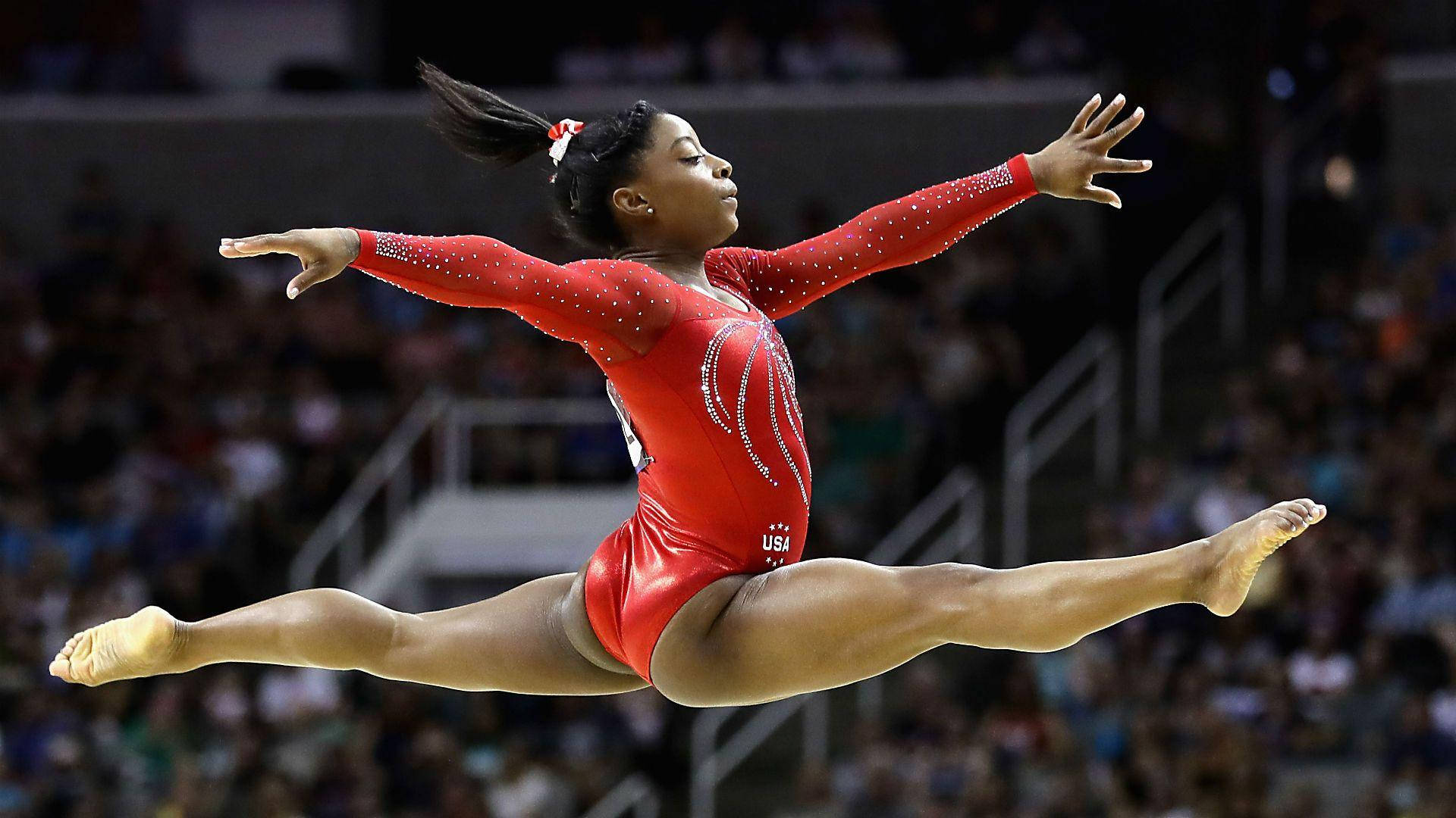 Gymnast 桌布 壁紙 — 一名身穿紅色緊身衣的女體操運動員在擠滿觀眾的場館中進行競技表演時，在空中完美地完成了一個分腿跳躍。
